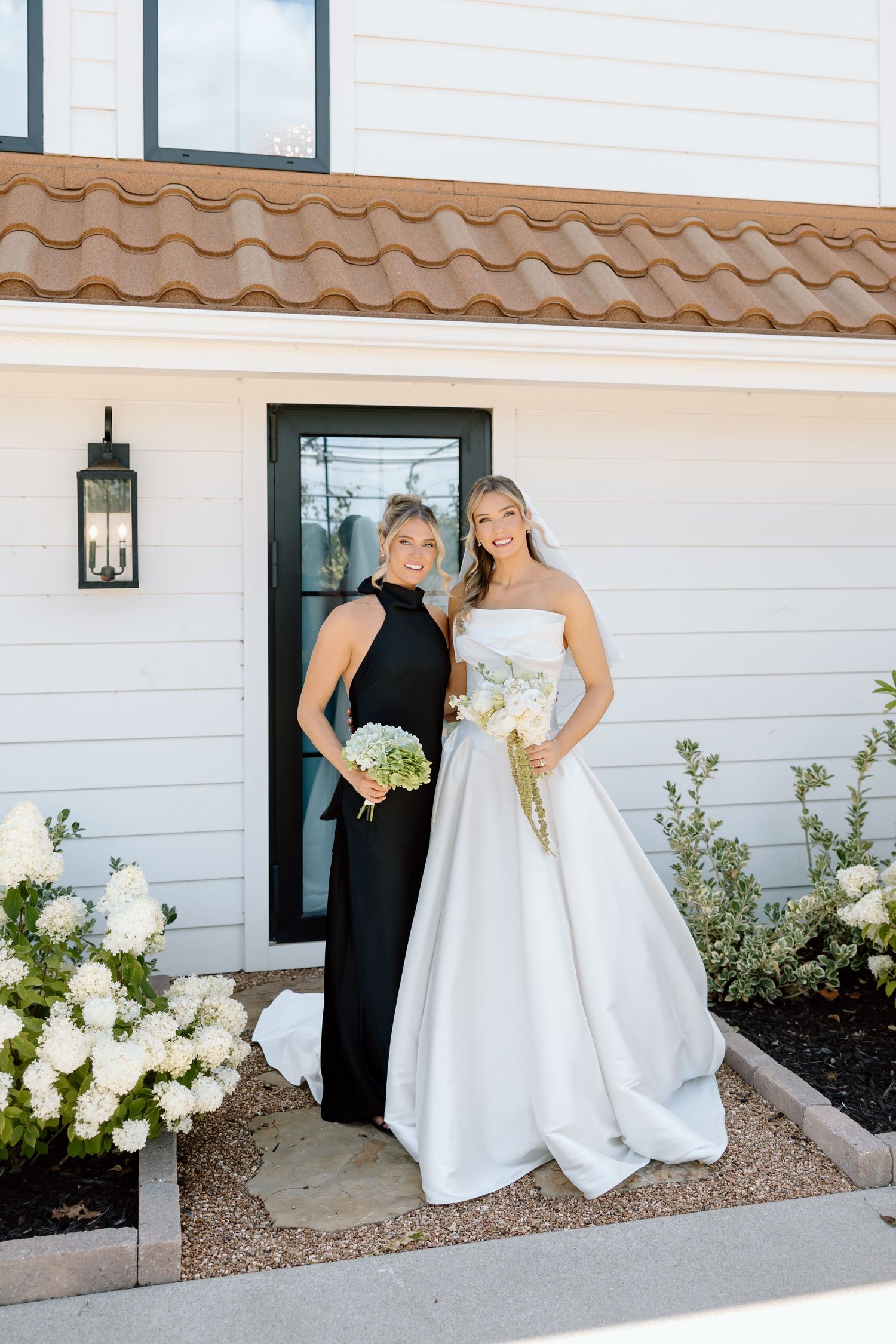 Bride in white strapless gown and bridesmaid in black halter dress pose with bouquets outside white home.
