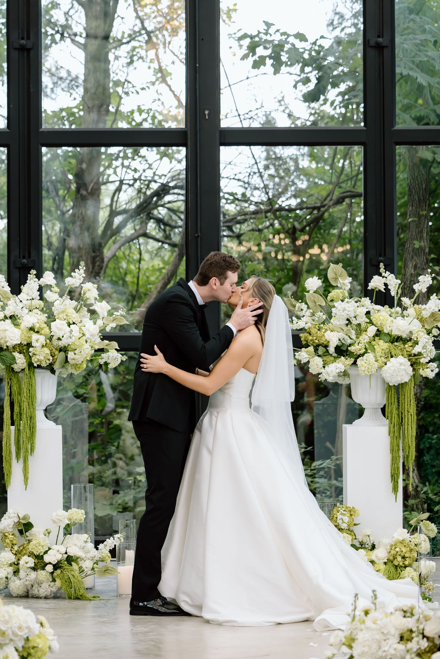Bride and groom share first kiss at altar surrounded by white floral arrangements and green hanging amaranthus in elegant venue.