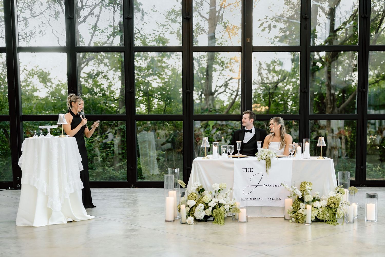 Elegant wedding reception with bride and groom at sweetheart table while a woman gives a toast near a white podium.
