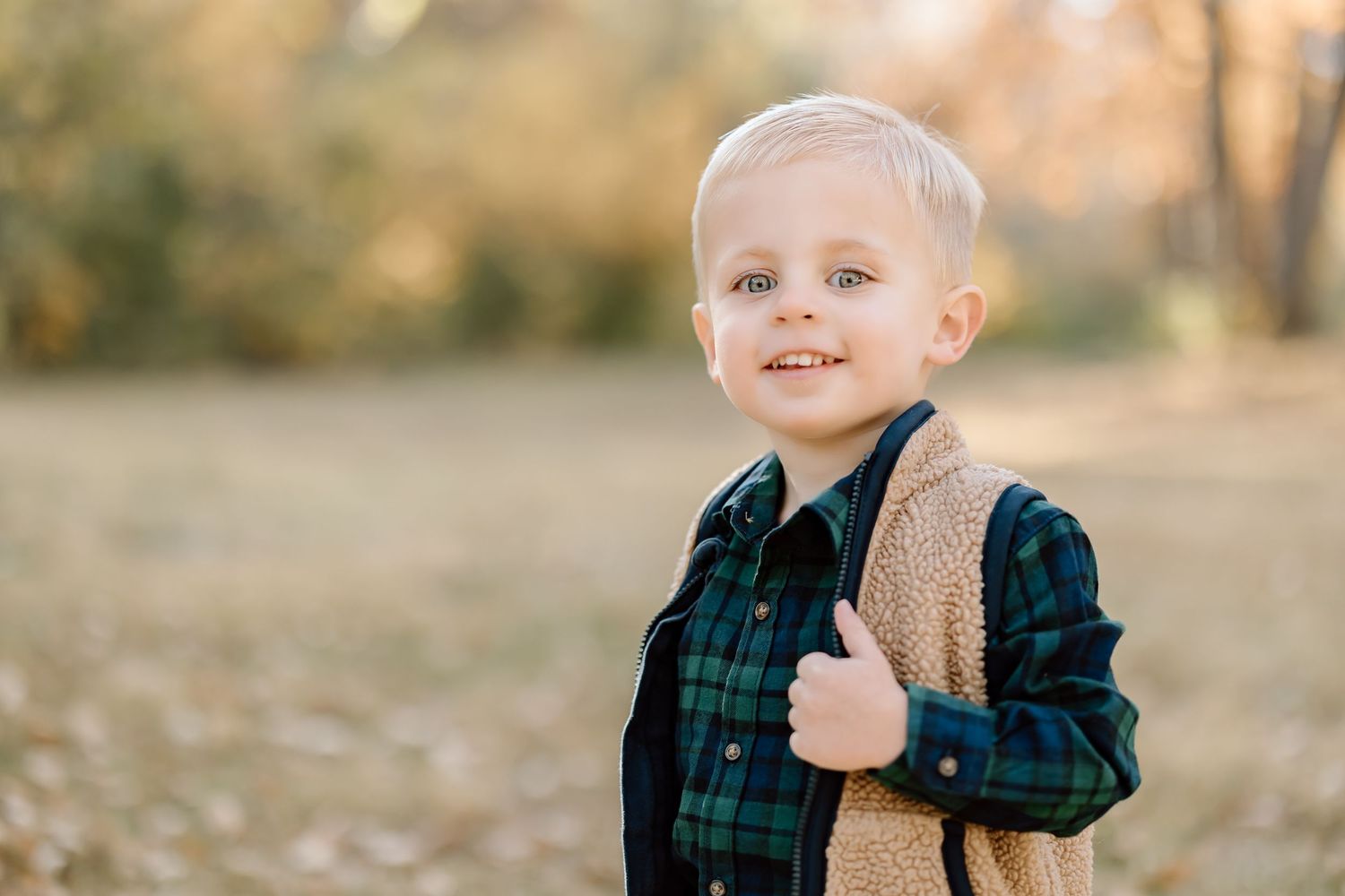 Child wearing green plaid shirt and sherpa vest smiles brightly in autumn setting.