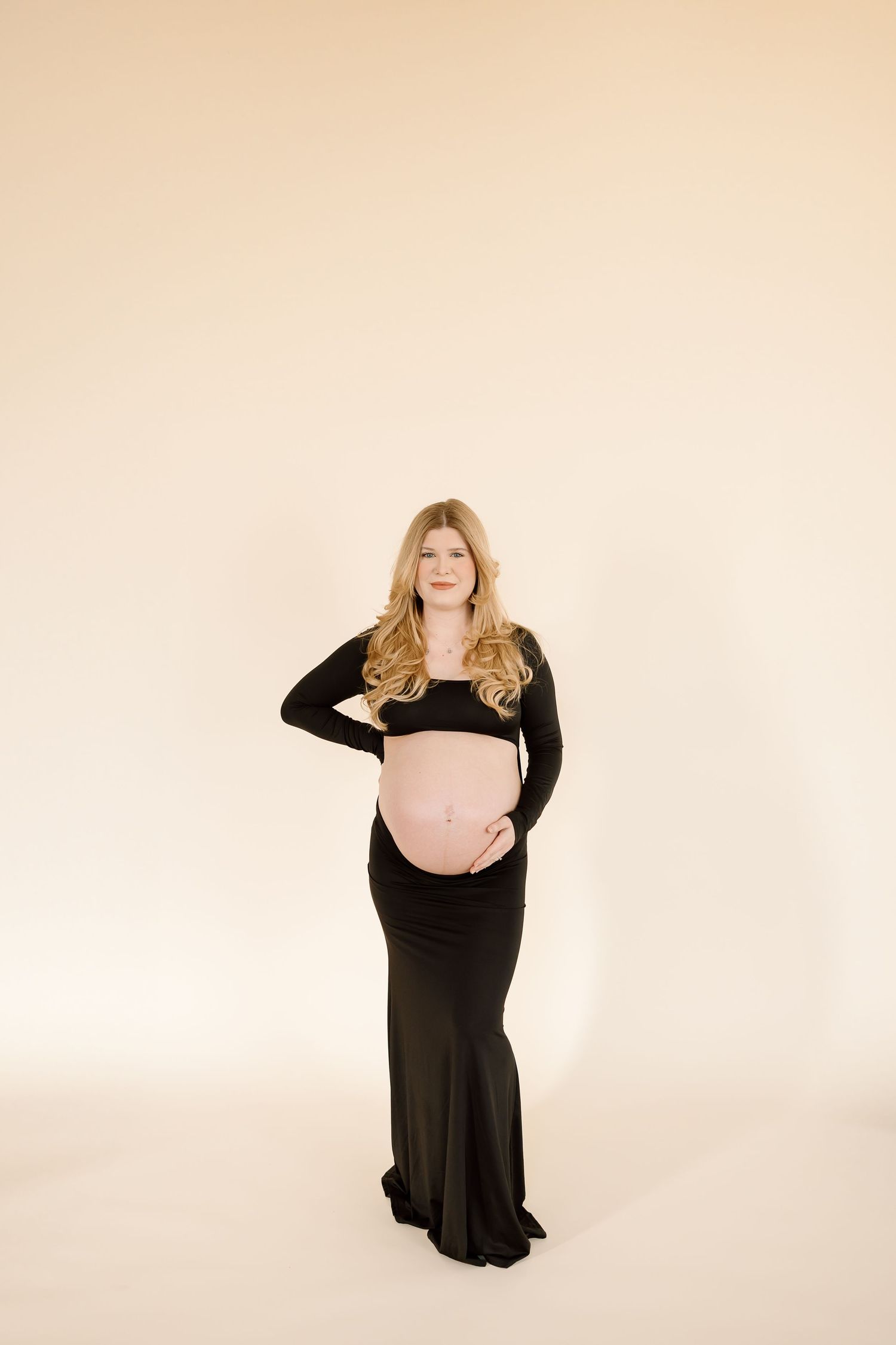 Stylish maternity portrait in black dress against beige background showcasing baby bump.