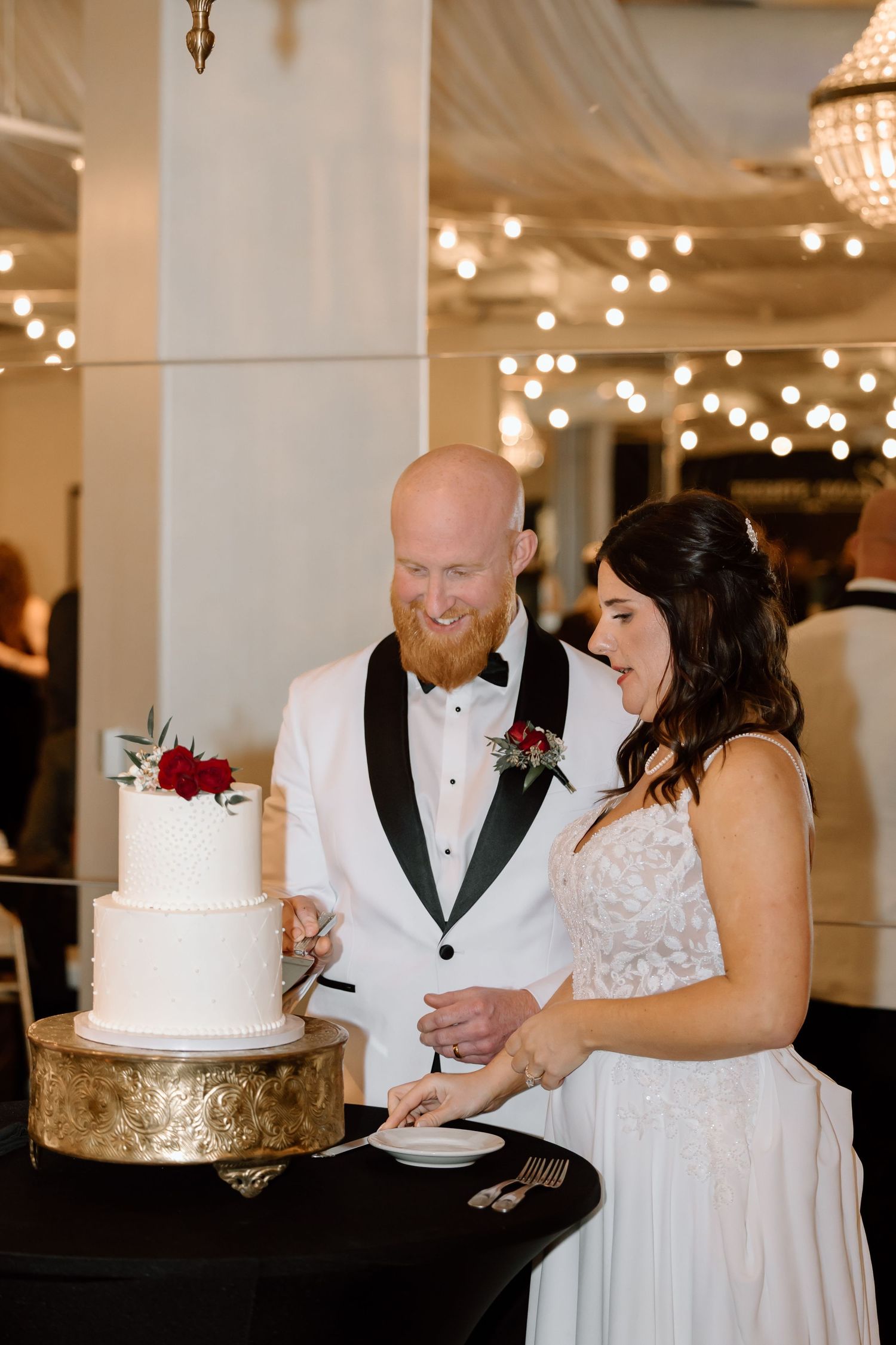Newlyweds share a romantic moment while cutting their elegant white and gold wedding cake at an indoor reception.