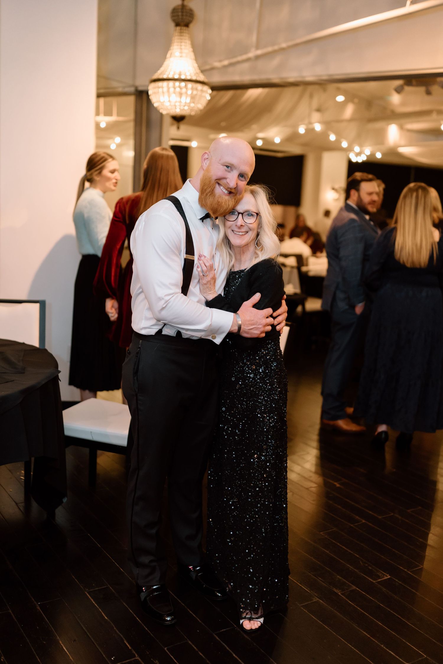 Couple dancing together on a dark wooden floor at an elegant indoor venue with chandelier and string lights.