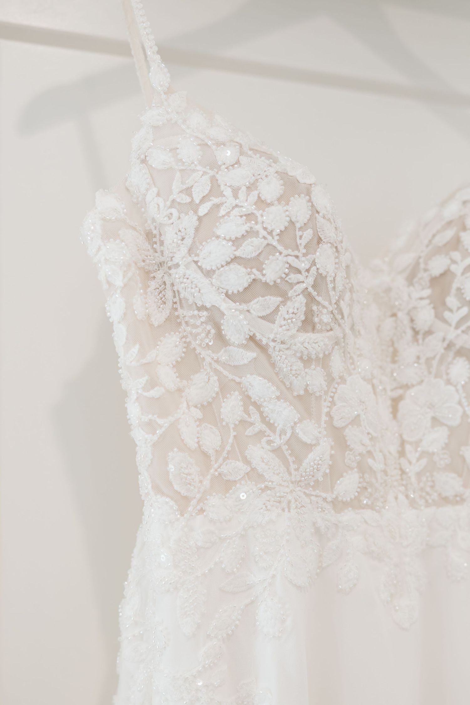 Delicate white lace and beaded details adorn the bodice of a wedding dress against a light background.