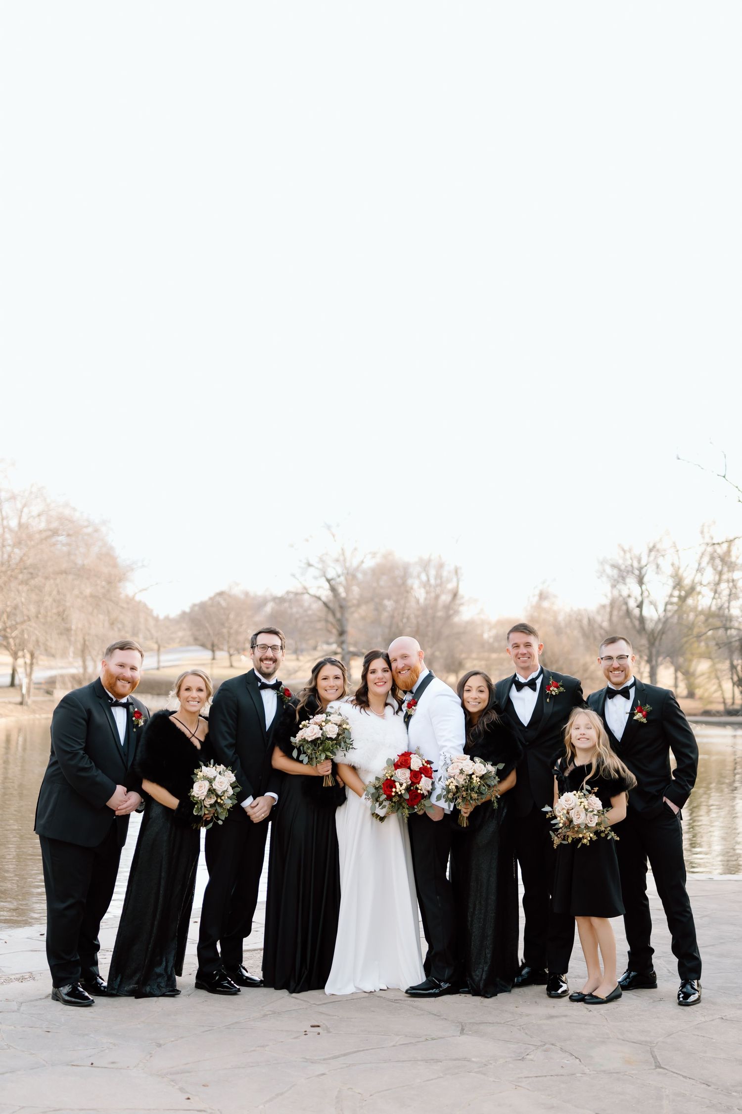 Wedding party in black and white formal attire poses together outdoors near a lake during a winter celebration.