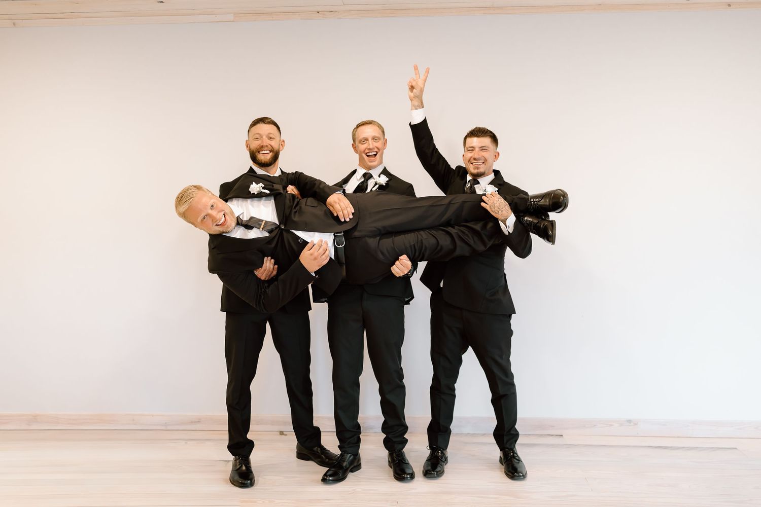 Four figures in black suits perform a synchronized dance move against a white wall.