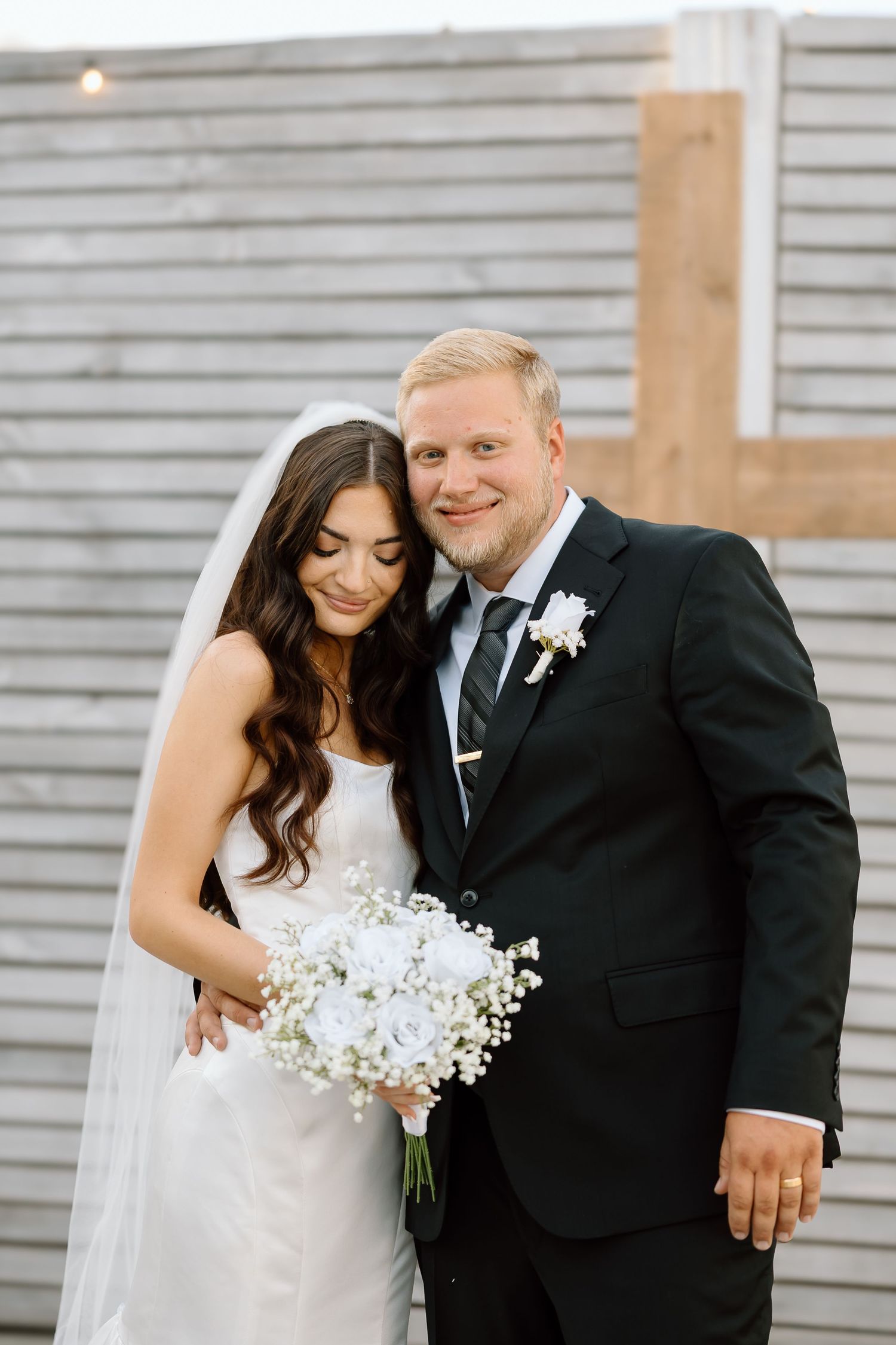 A couple shares intimate wedding moments in front of a gray siding wall while holding a white floral bouquet.