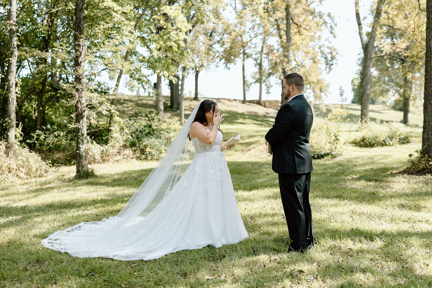 A couple shares a romantic moment in their wedding attire on a sunny lawn surrounded by trees.