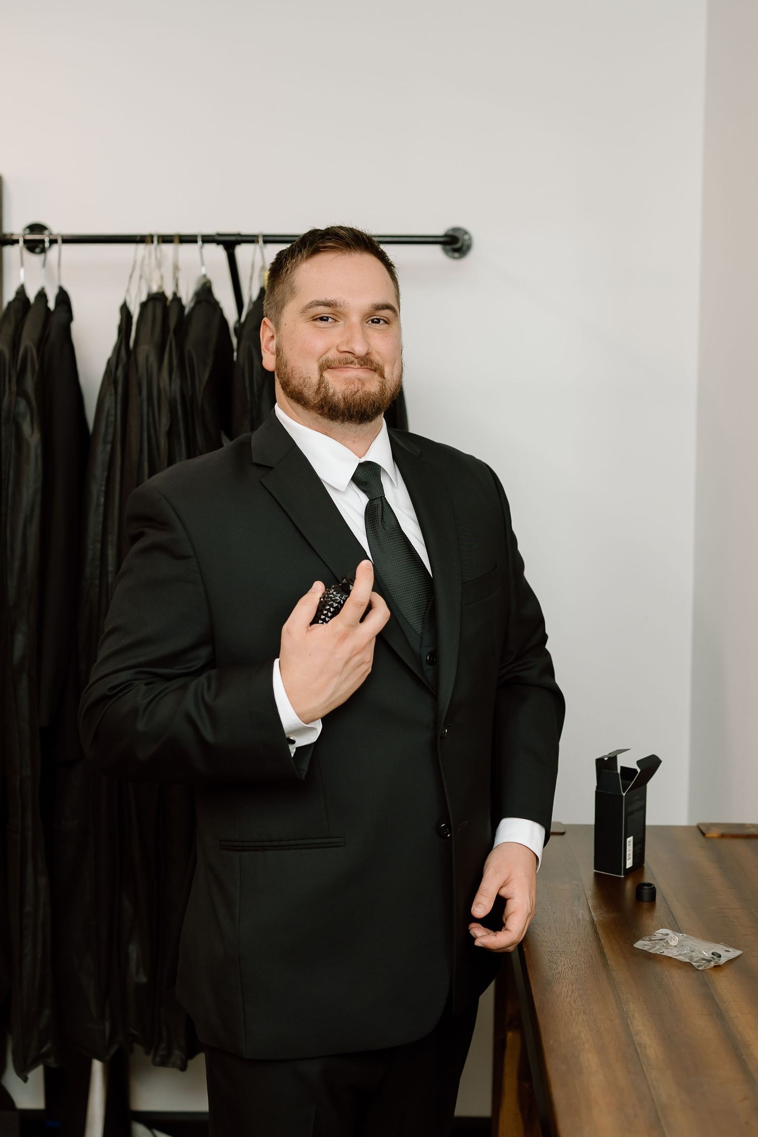 A person in a black suit and tie stands in front of a clothing rack while adjusting their jacket.