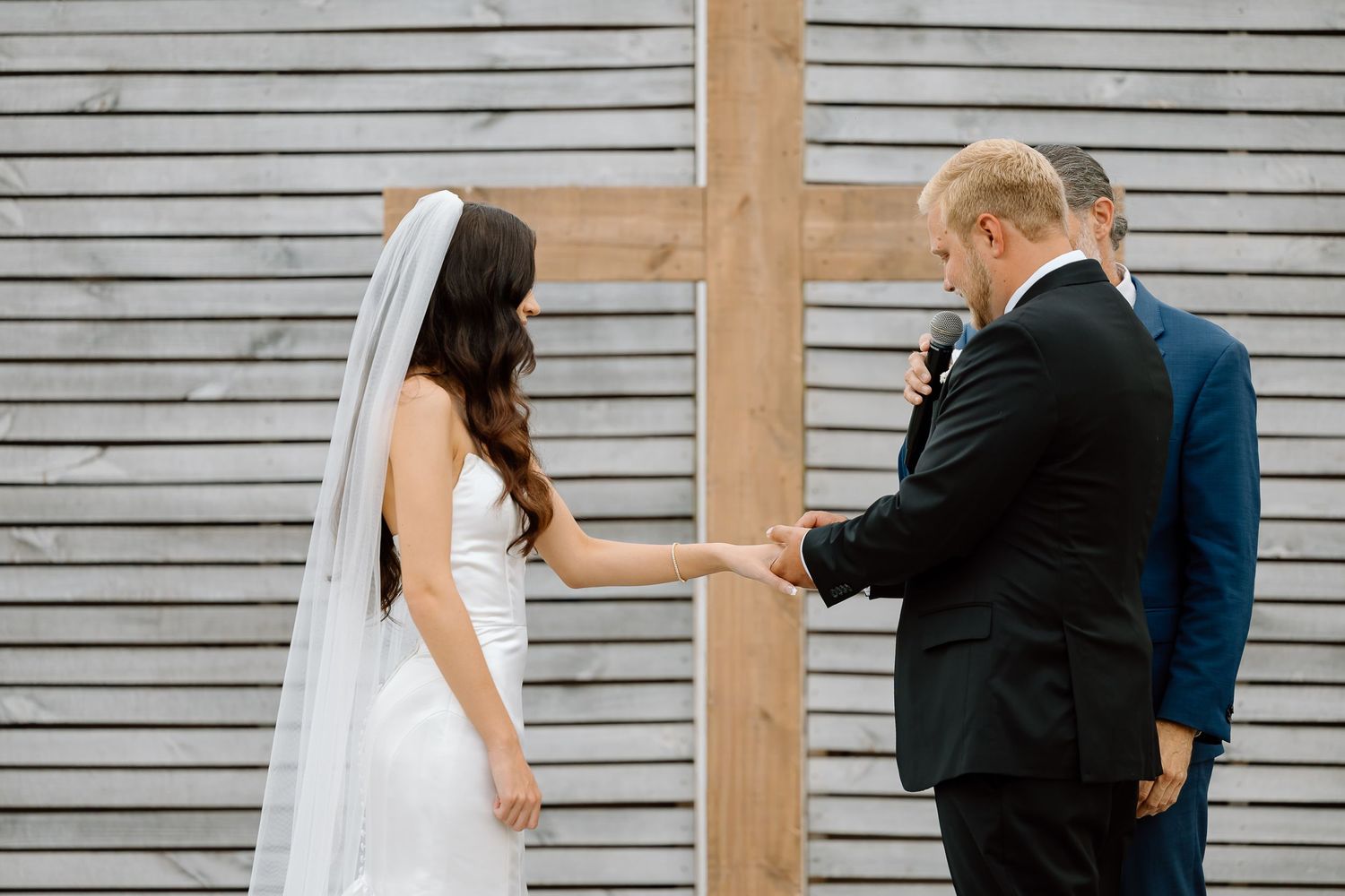 A wedding couple poses for photos in front of a gray slatted wall with natural light filtering through wooden beams.