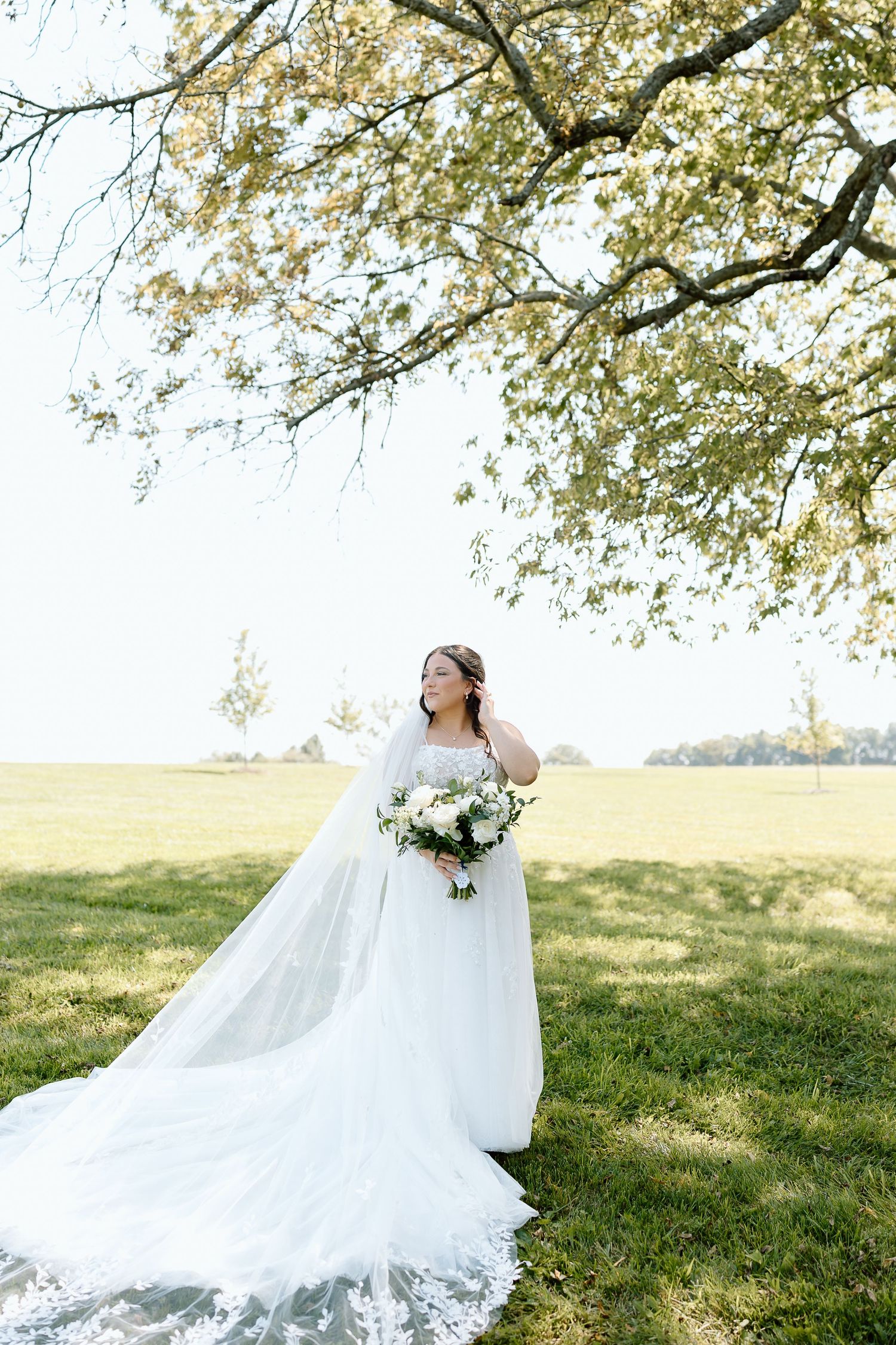 Bride in flowing white wedding dress twirls gracefully on grassy hillside under blooming spring trees.