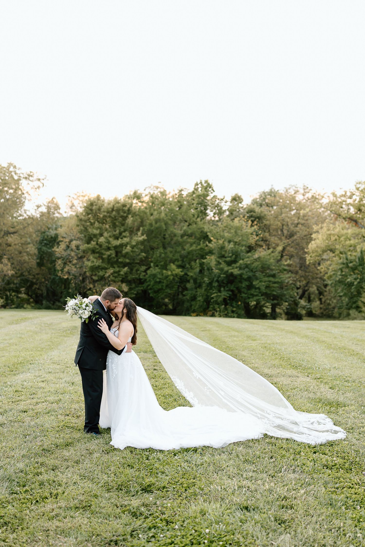A romantic wedding couple embraces in a sunlit meadow as the bride's long veil flows gracefully in the breeze.