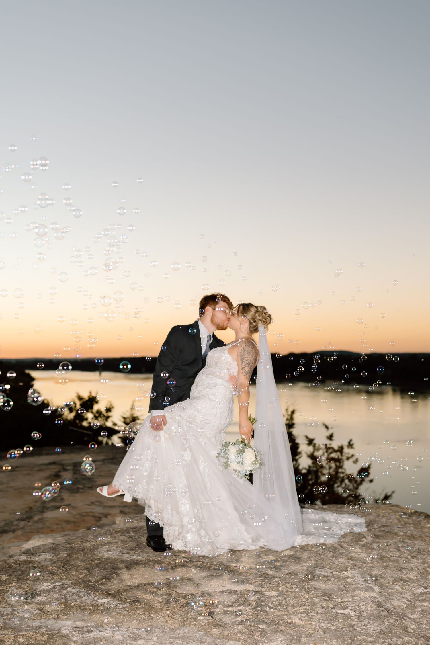 A couple in wedding attire share a romantic moment at sunset by a lake with bubbles floating in the air.