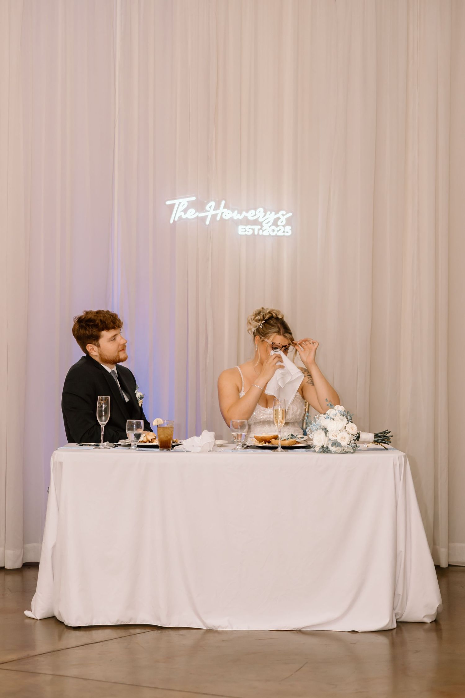 Wedding party seated at long white-clothed head table in elegant reception venue with draped backdrop.