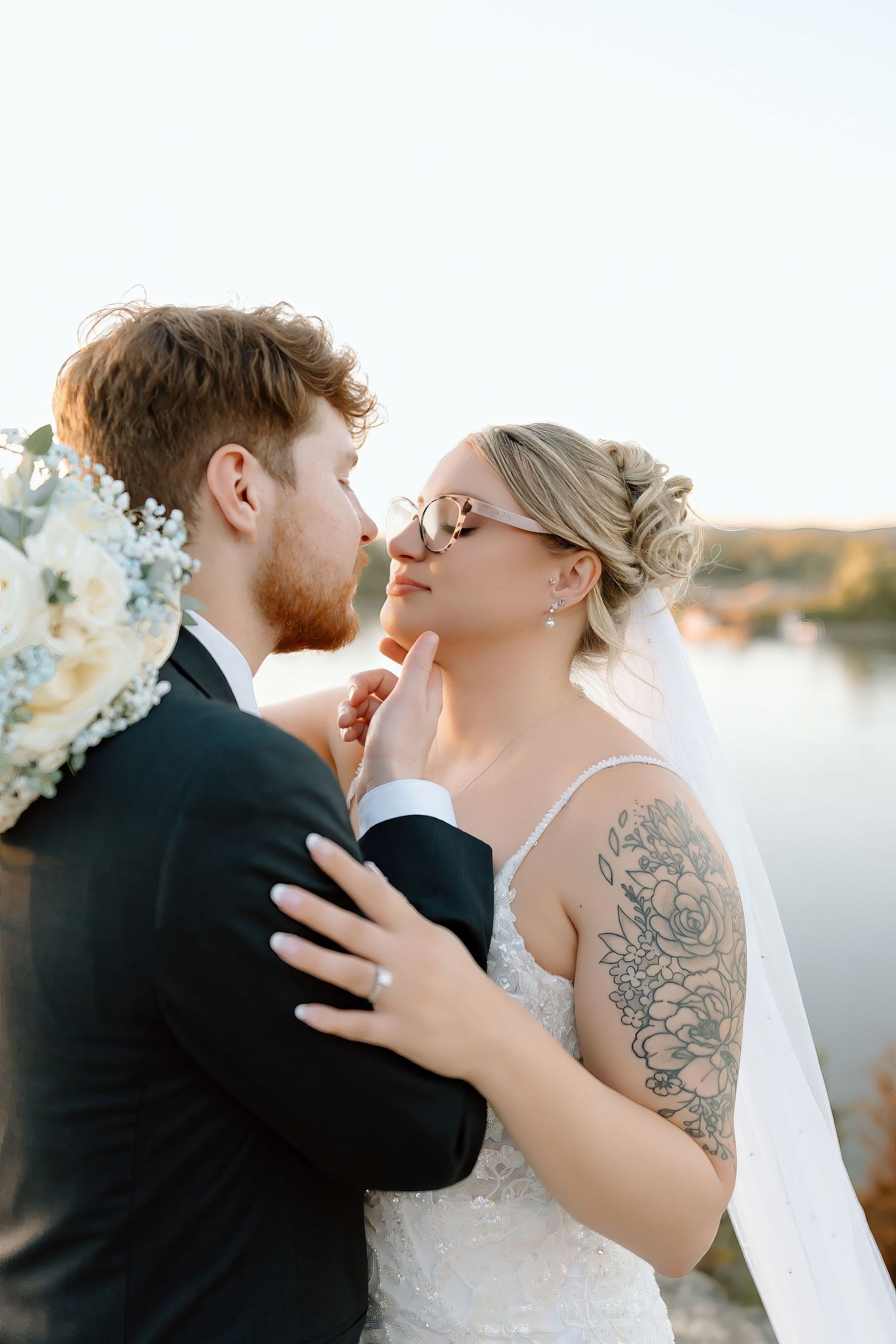 A romantic wedding couple embraces during sunset by a lake, with the bride wearing a white dress and showing a shoulder tattoo.