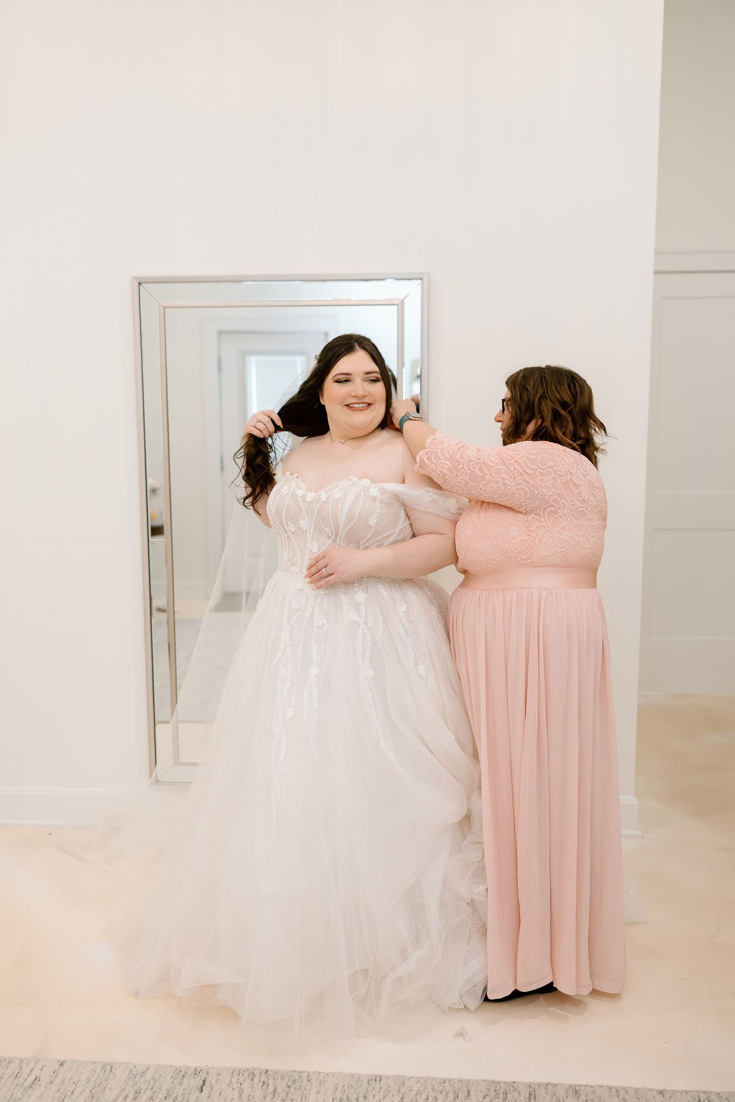 Two people in blush pink and white formal dresses embrace in a bright studio setting.