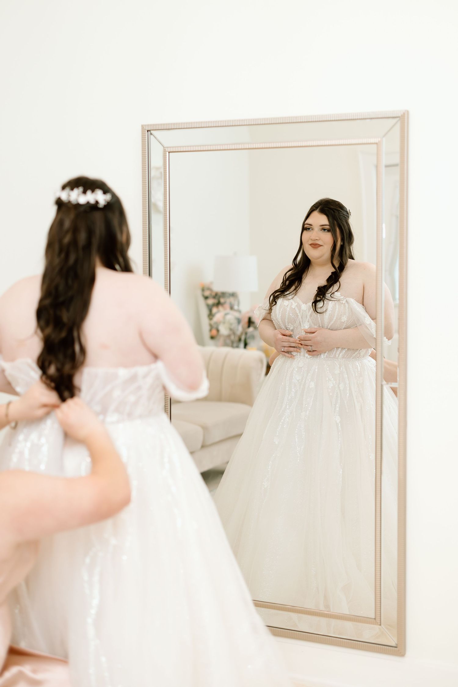 A bride in a white wedding gown looks at her reflection in three different mirror views.