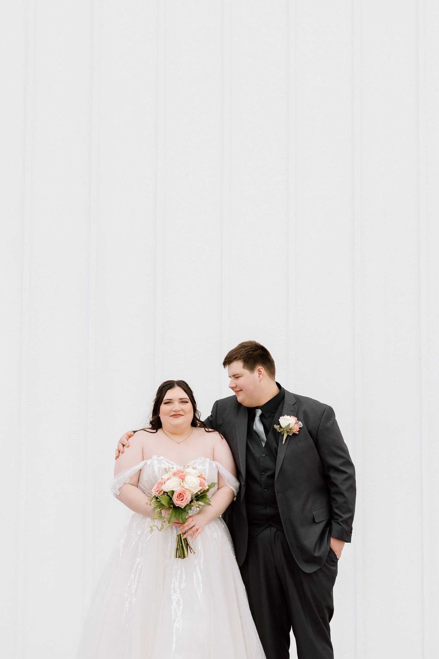 A couple in wedding attire poses against a white backdrop with a bouquet of peach and coral flowers.