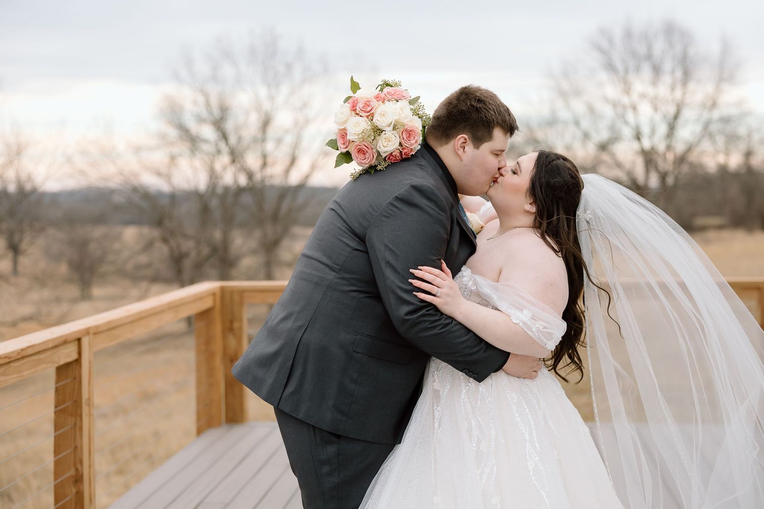 A couple shares a romantic kiss on a wooden deck with a bouquet of pink roses and flowing veil in the winter landscape.