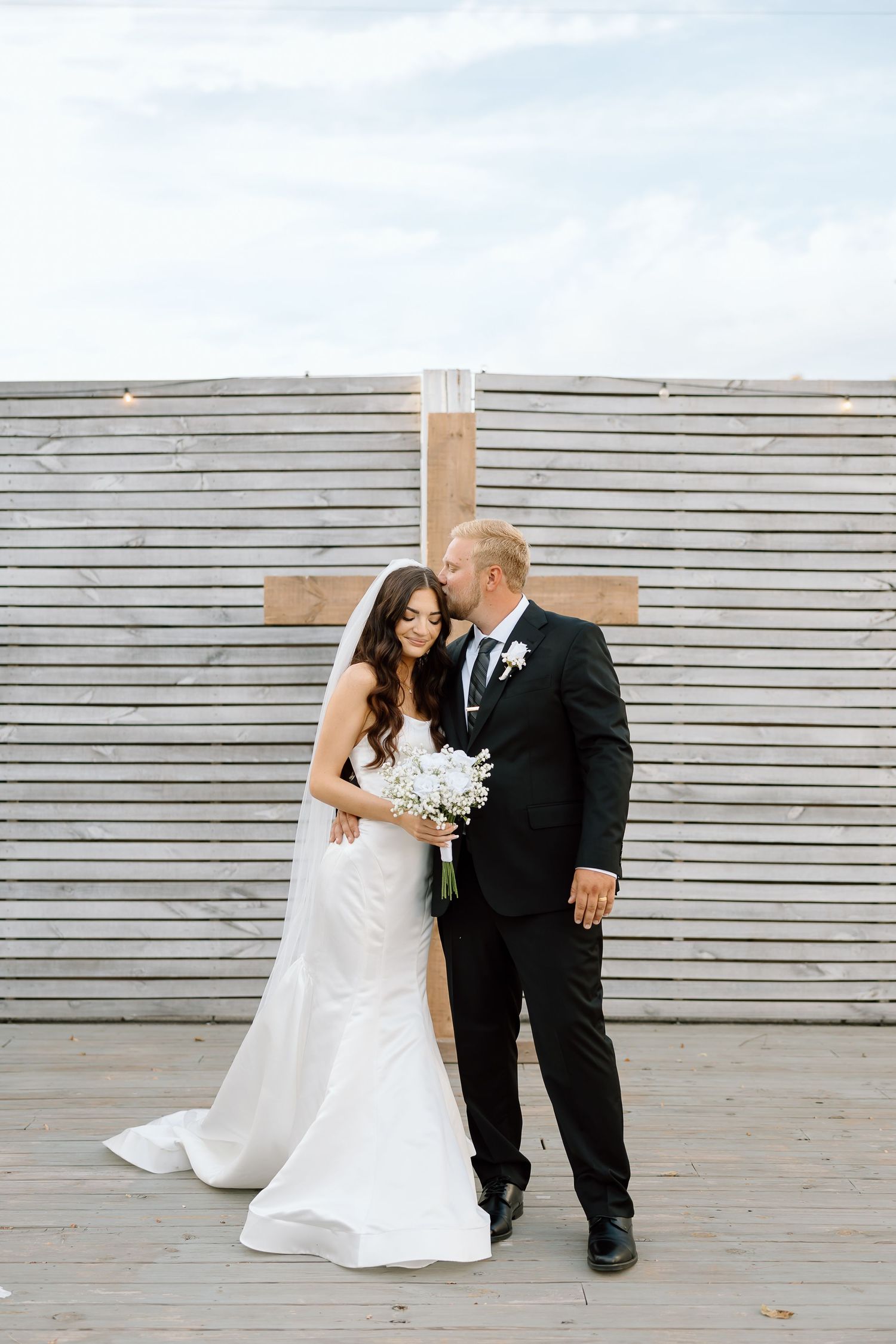 A couple in wedding attire poses against a modern gray wall during an outdoor photoshoot.