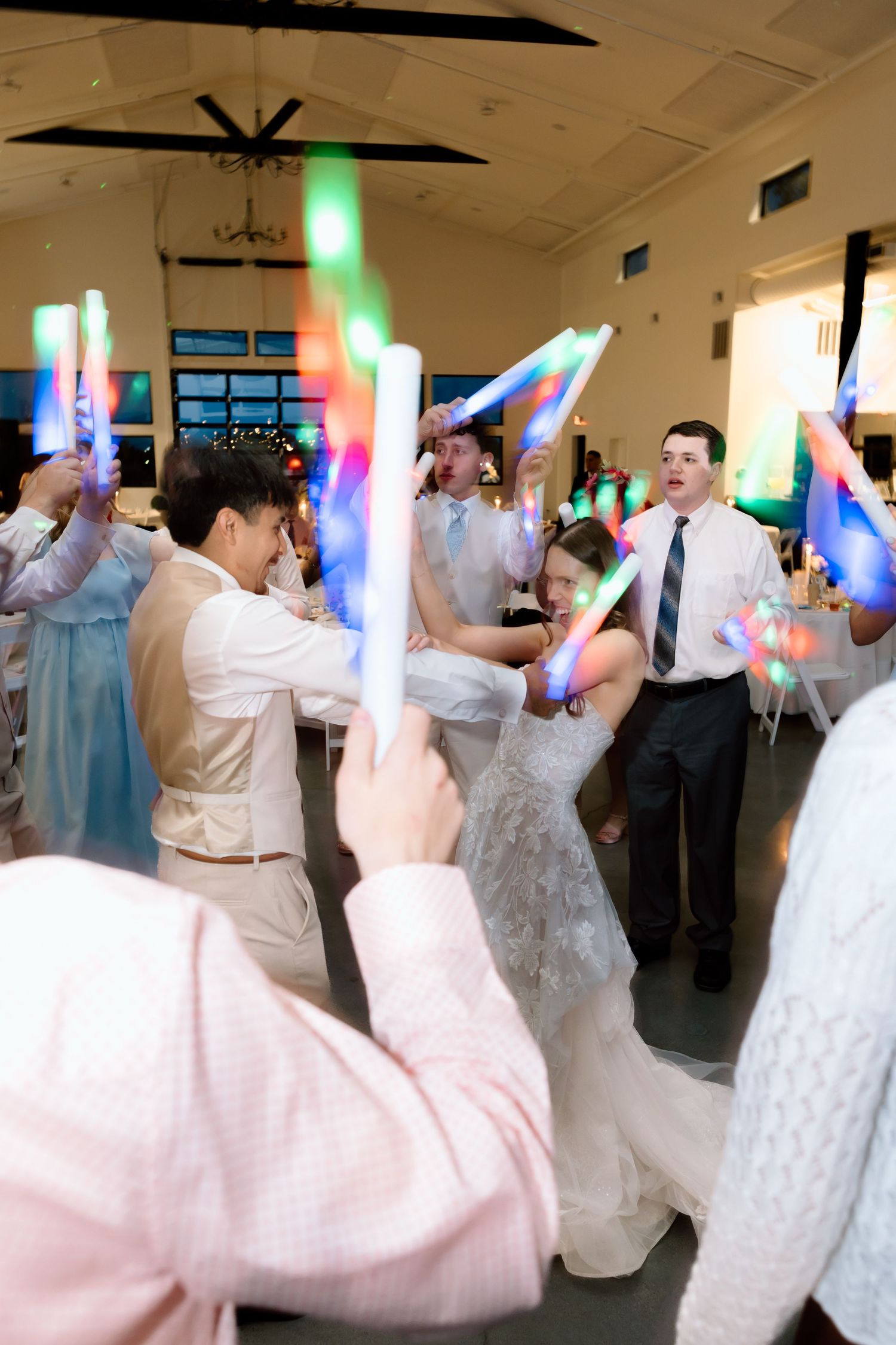 Bride and groom dancing with guests holding colorful LED foam glow sticks at wedding reception.
