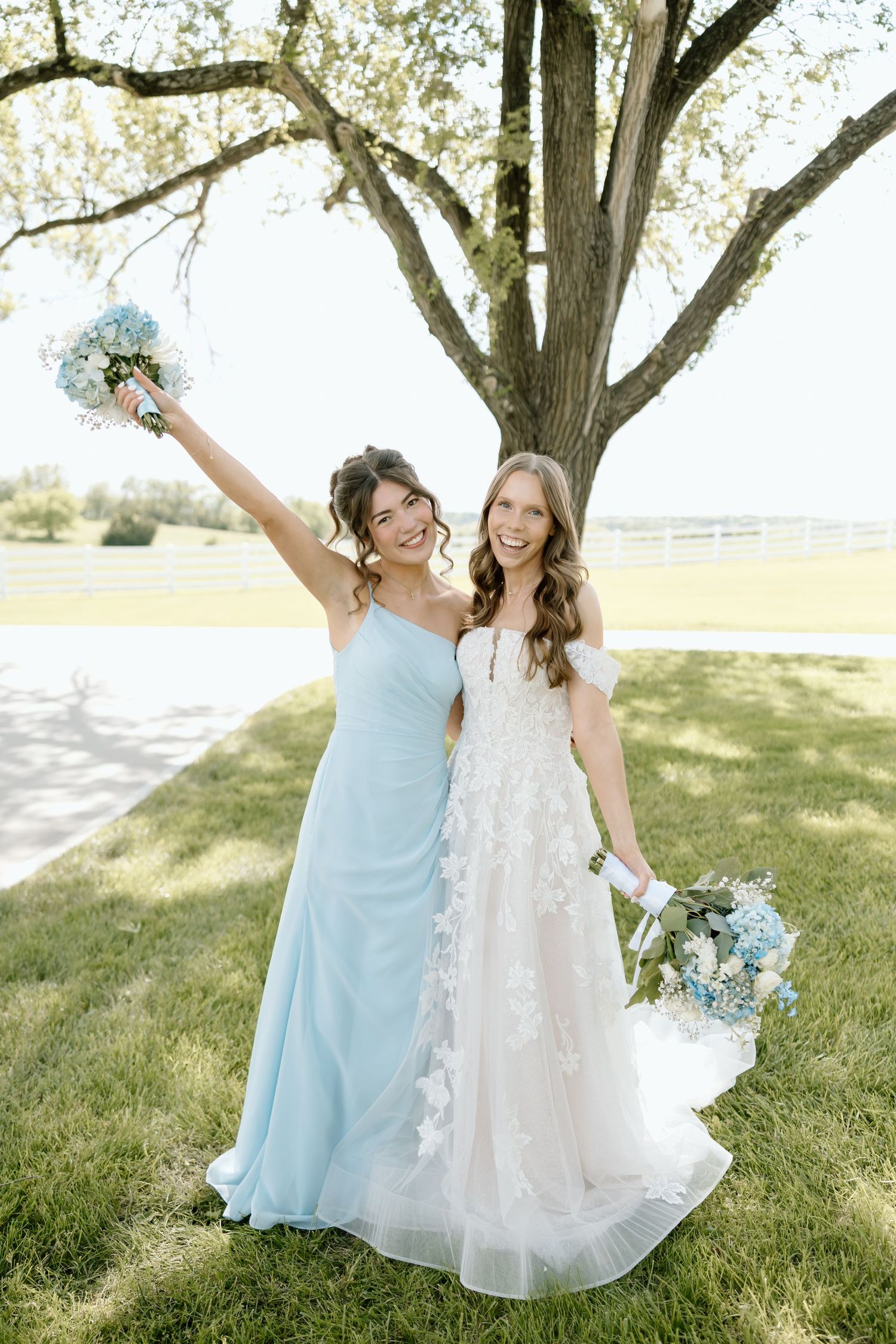 Bride in white lace gown and bridesmaid in light blue dress holding bouquets outdoors under a tree.