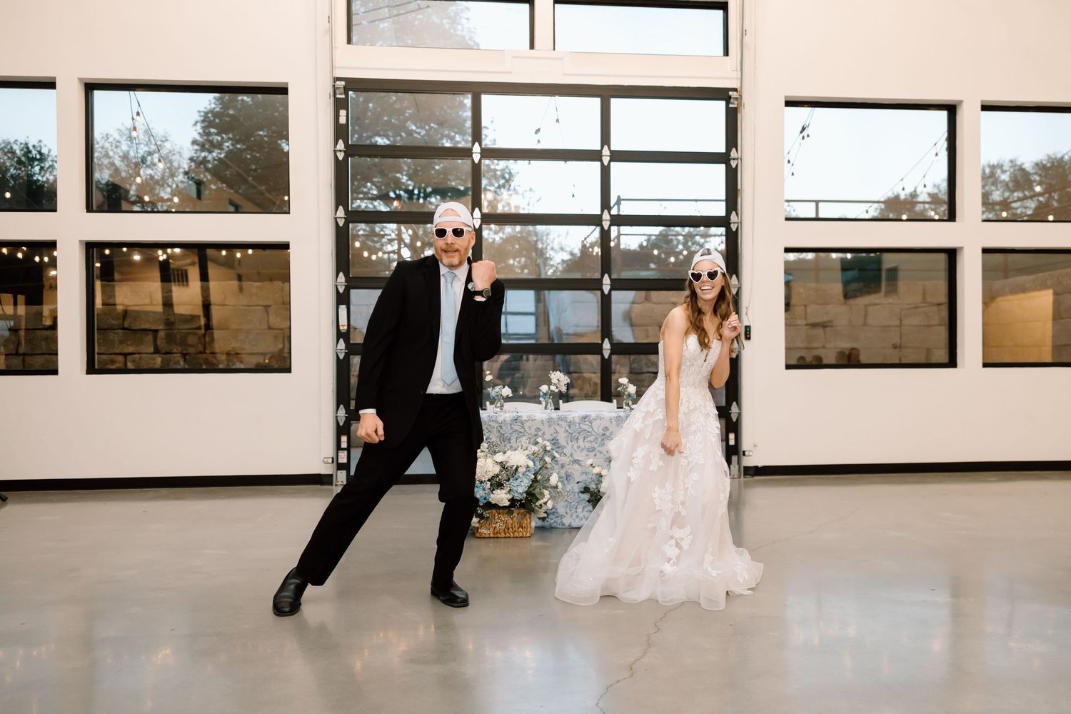 Bride in ruffled gown and groom in black suit wearing sunglasses dancing playfully in a modern wedding venue.