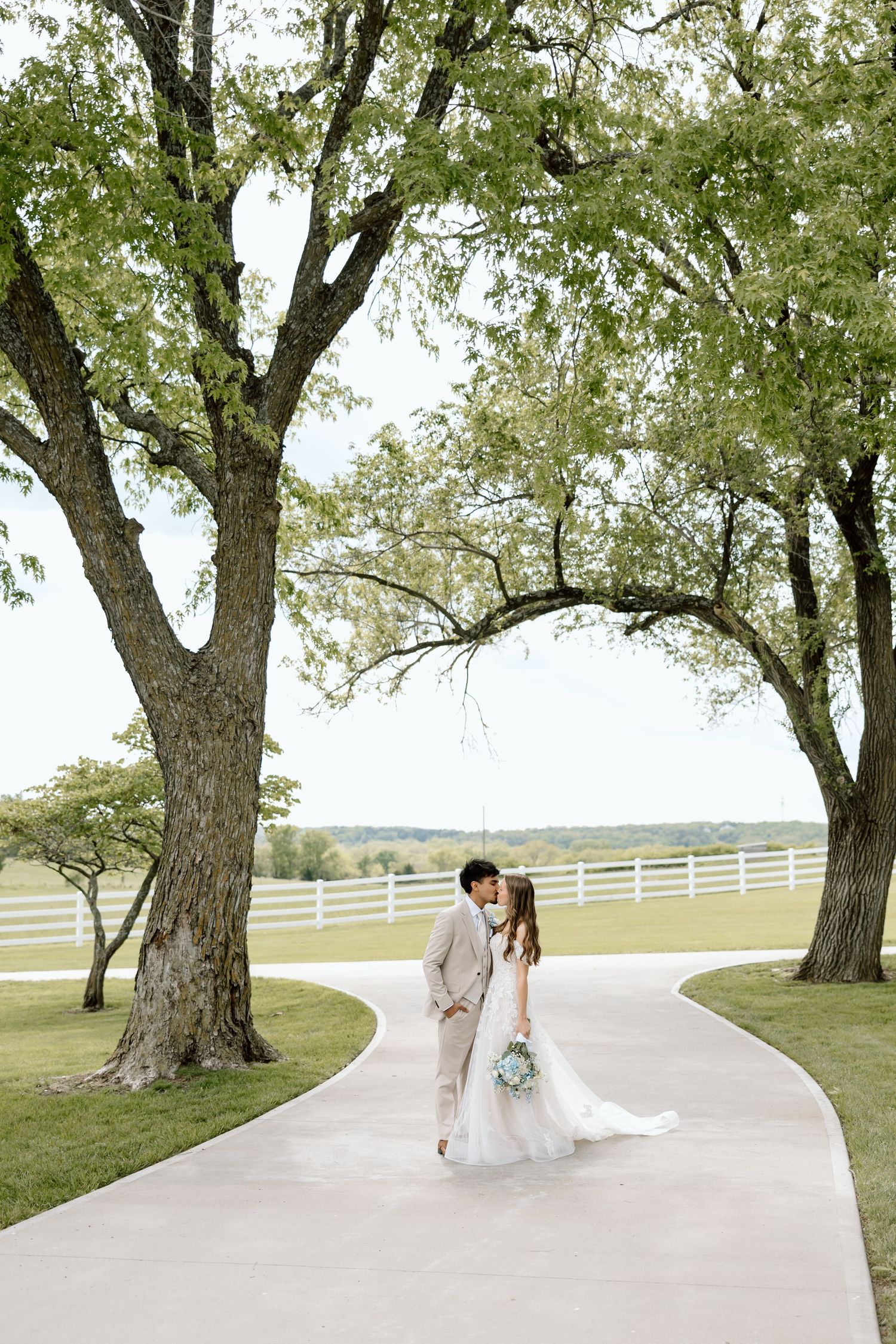 Bride and groom share a romantic kiss on a winding path framed by large trees at an outdoor wedding venue.