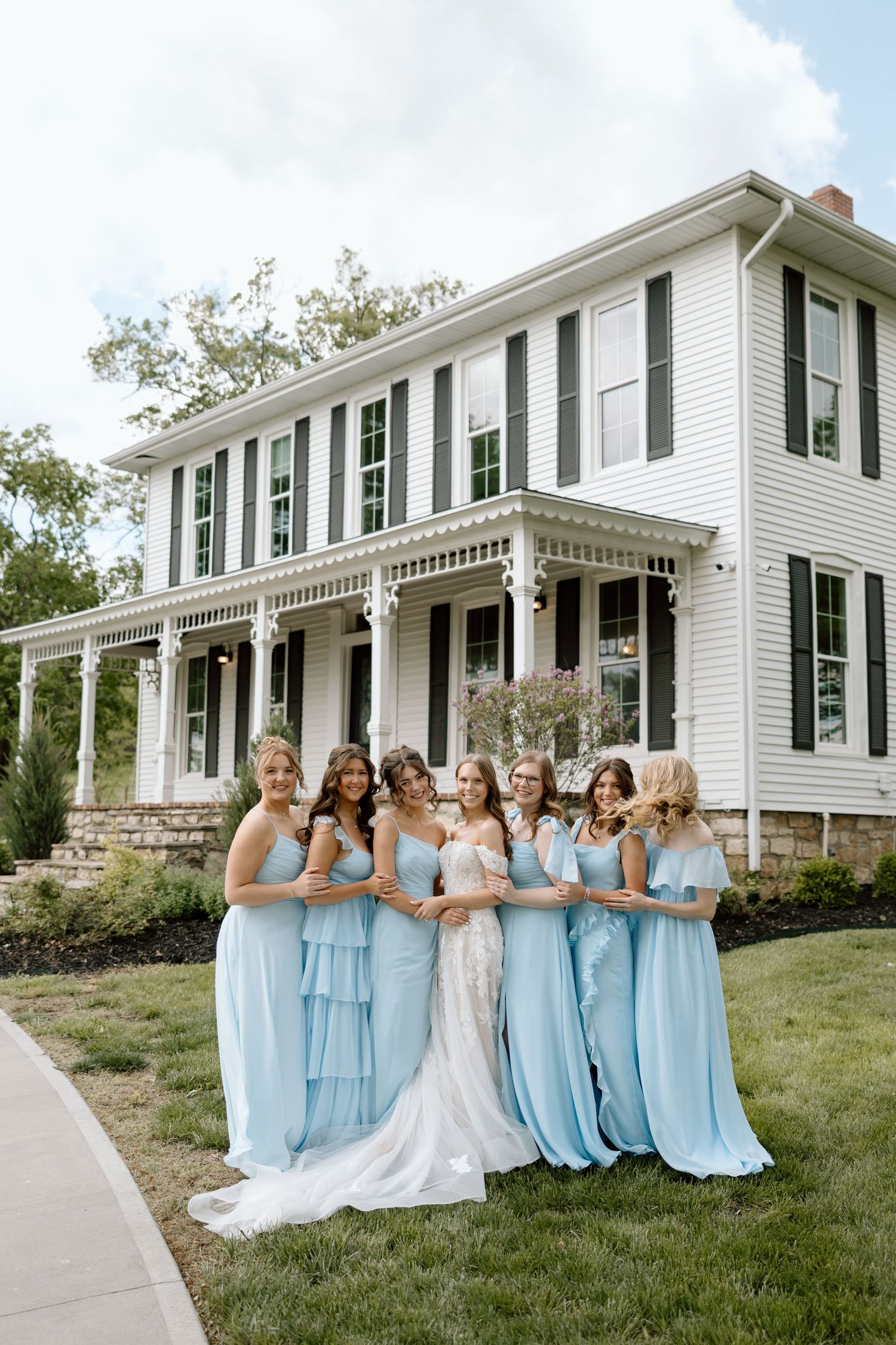 Bride in lace gown poses with bridesmaids in light blue dresses in front of a white colonial home.