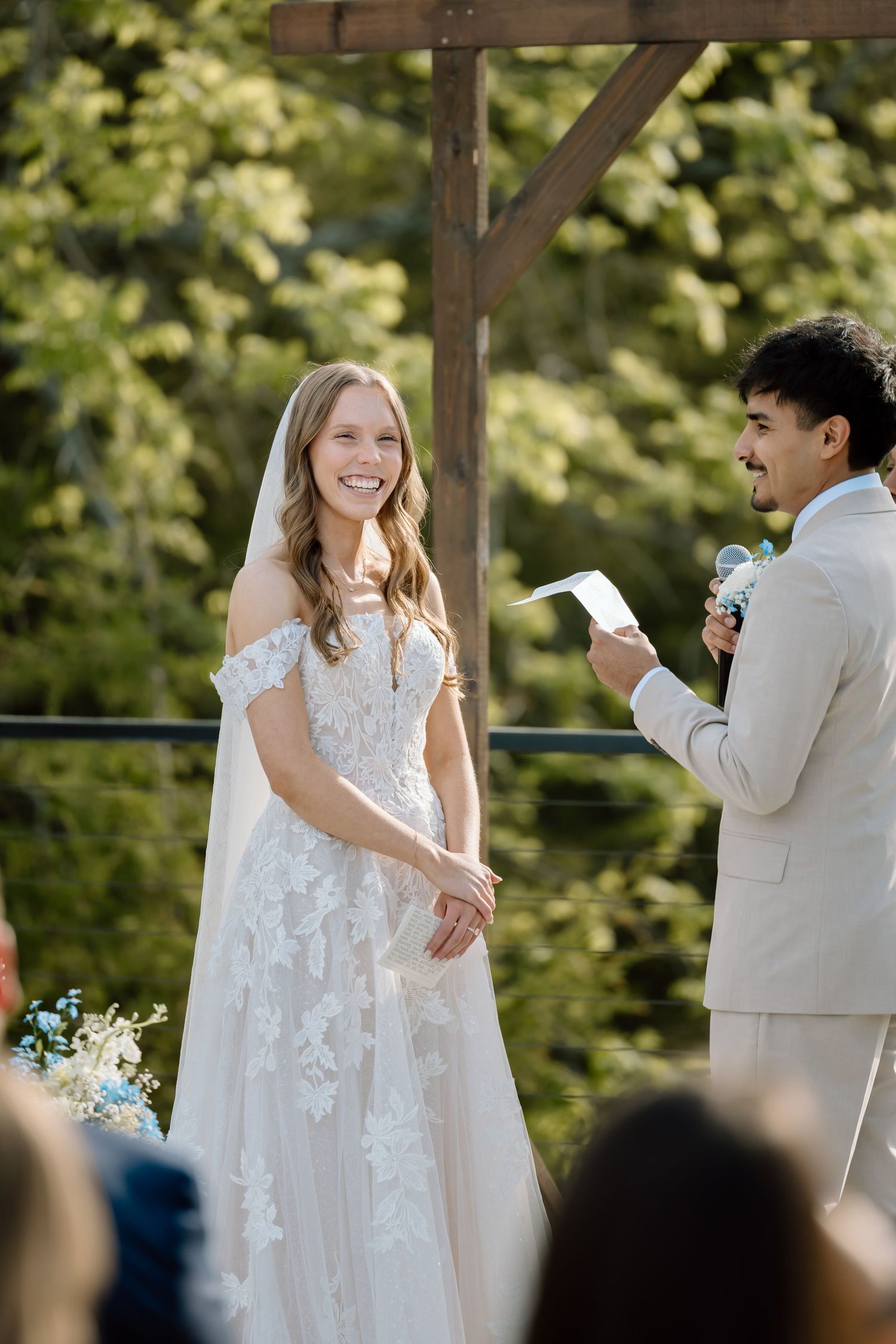 A smiling bride in a lace wedding gown listens as the groom reads vows at an outdoor ceremony.