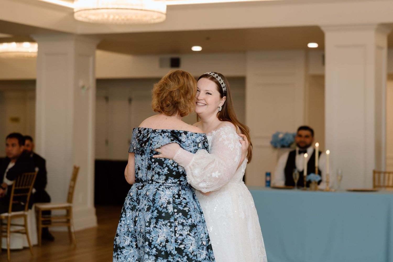 Bride in white lace gown embraces a woman in blue floral dress at a wedding reception venue.