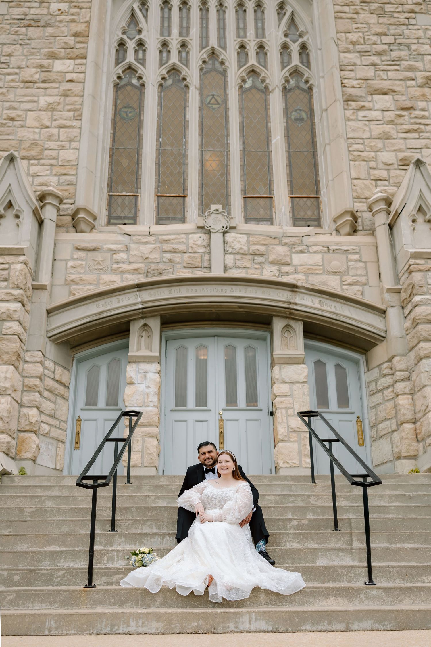 Bride and groom sitting on stone church steps, bride in flowing white gown, groom in black tuxedo.