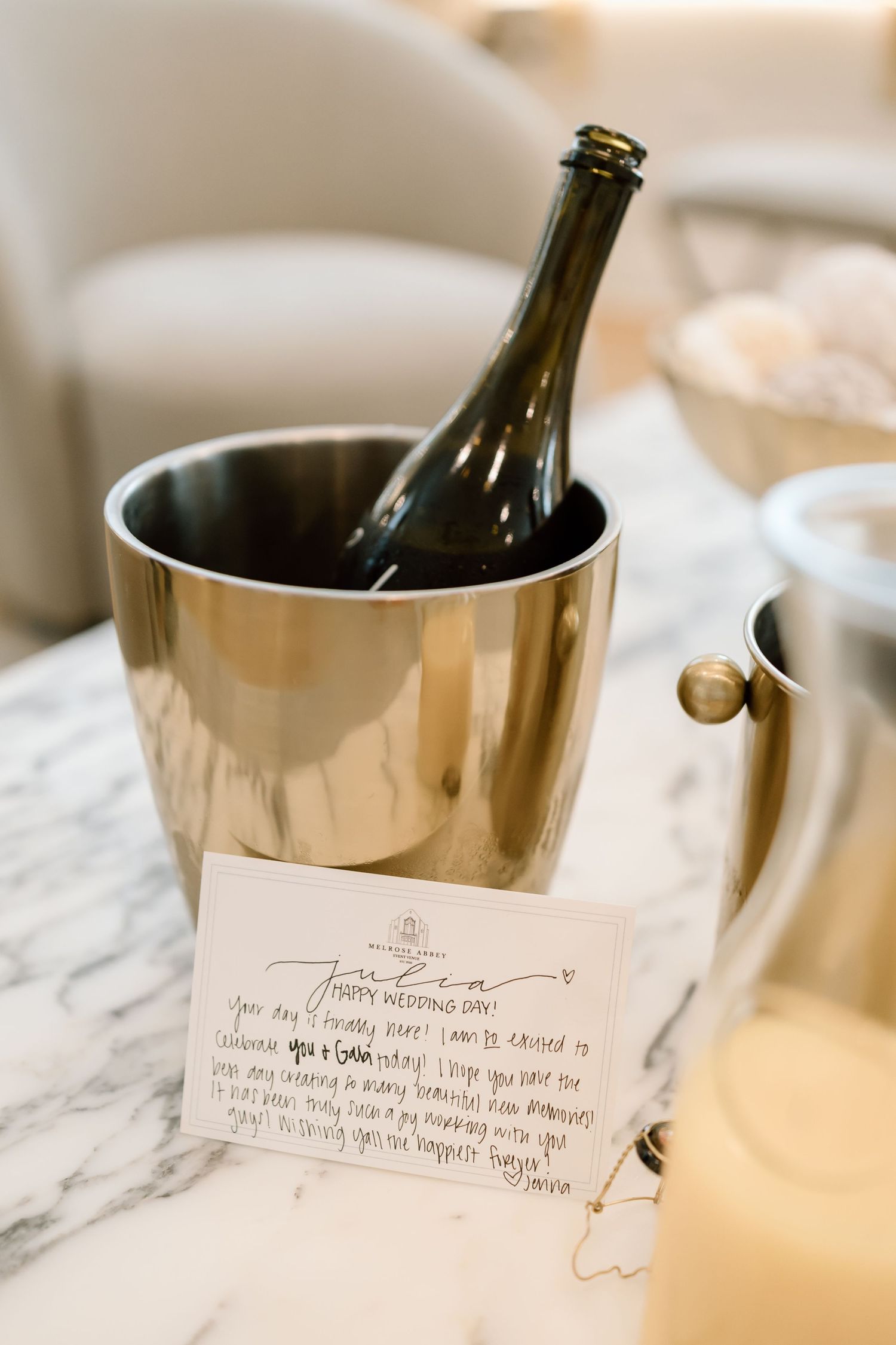 Silver champagne bucket with bottle and handwritten wedding day card on marble table.