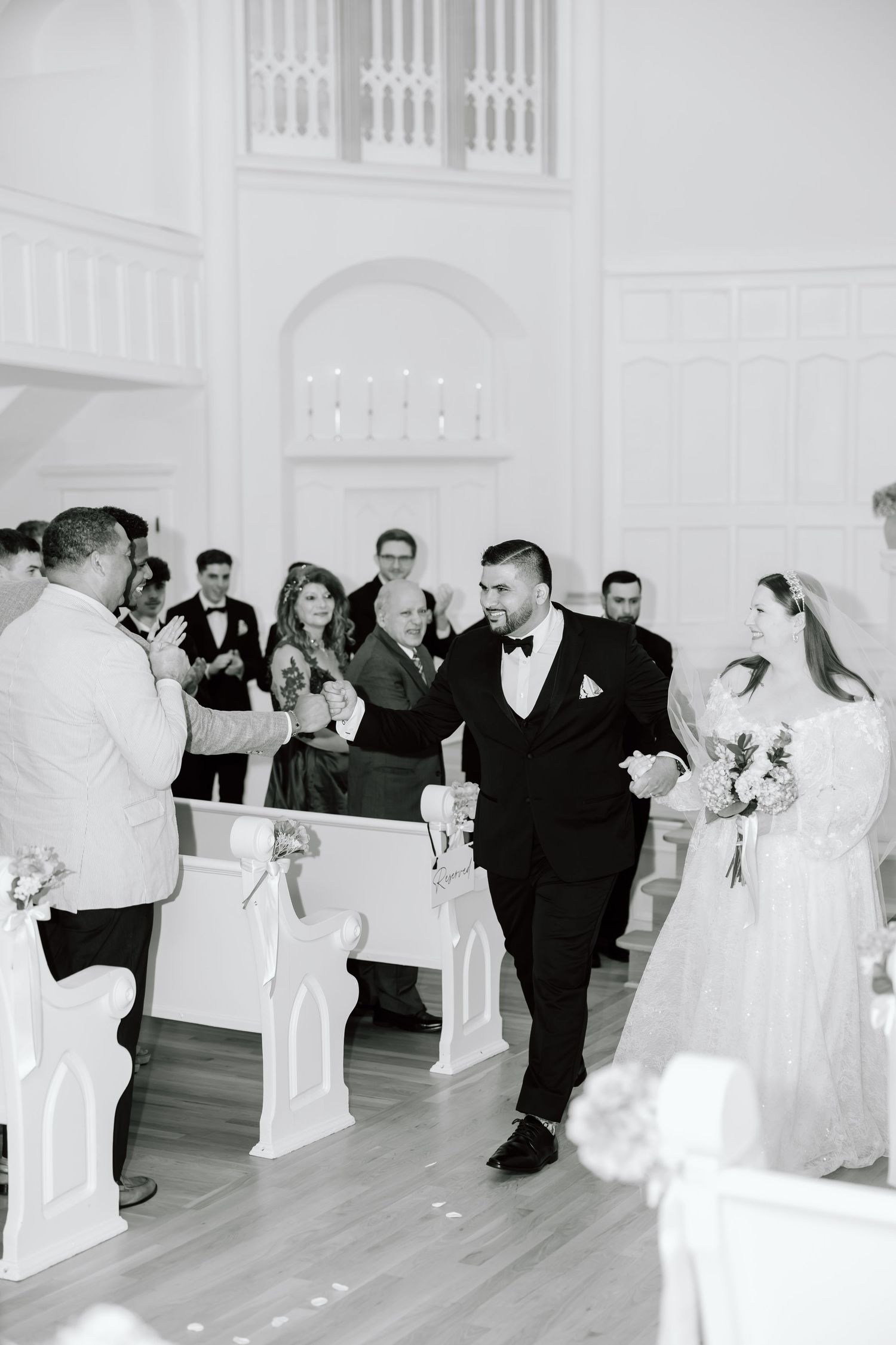 Black and white photo of a groom walking bride down church aisle during elegant wedding ceremony with guests applauding.