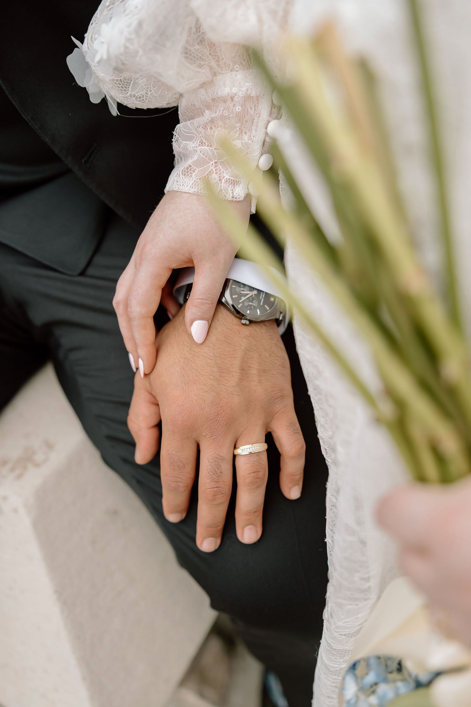 Bride and groom hands together showing wedding rings, with the groom wearing a watch and the bride in lace dress.