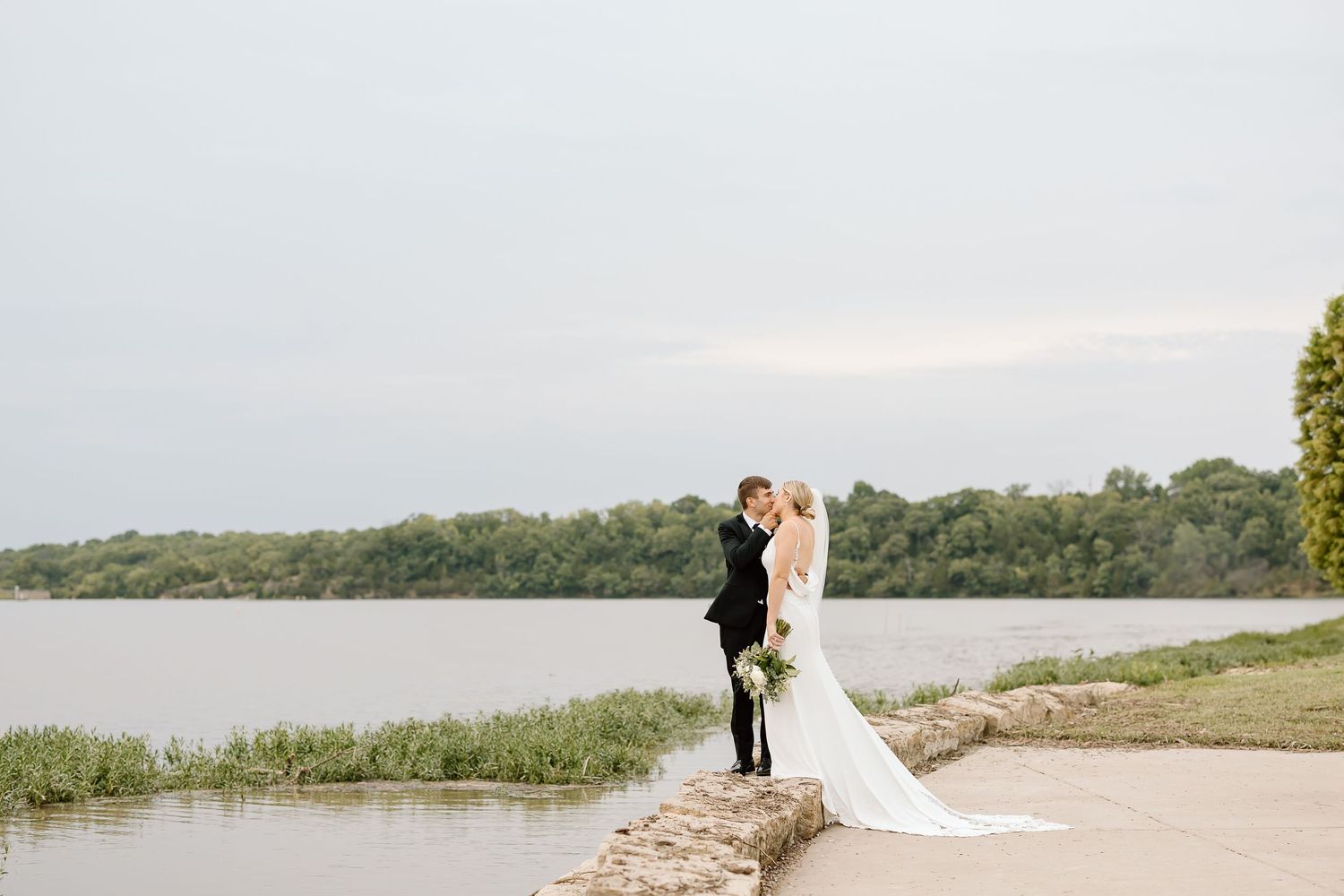 Couple in wedding attire embraces on a scenic lakeside path during golden hour against a lush green backdrop.