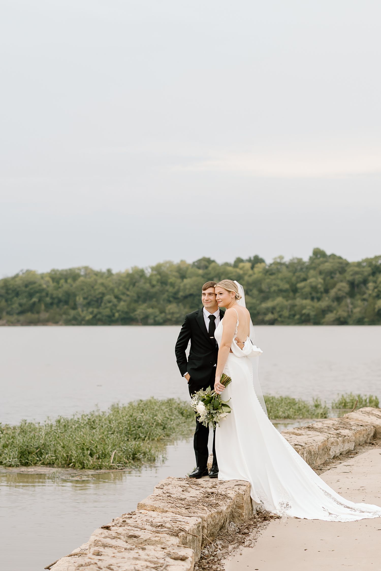A wedding couple stands on a stone wall overlooking a serene lake surrounded by trees on an overcast day.