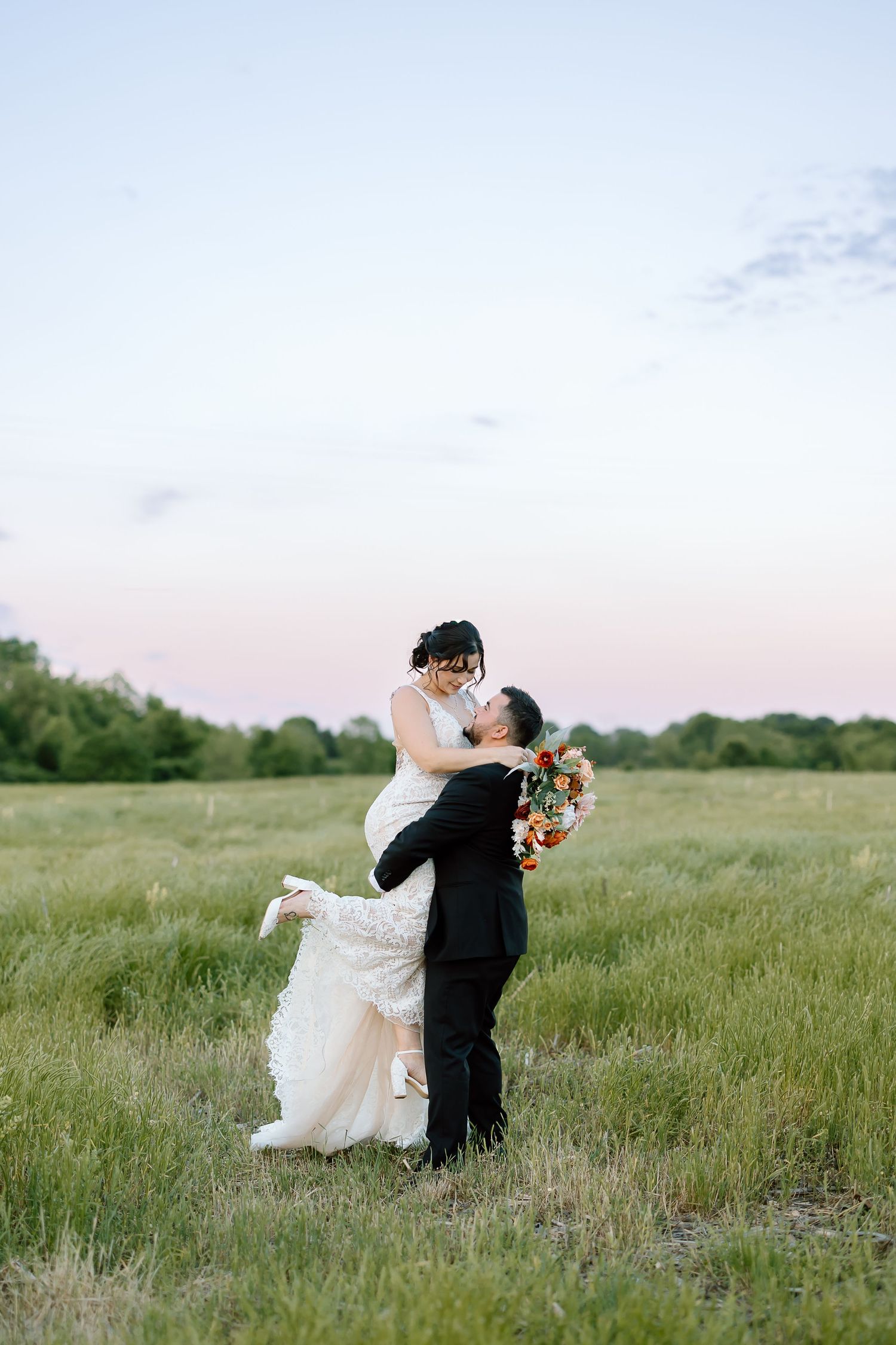 A couple shares a romantic dance together in a serene meadow during sunset, with the bride in a flowing white dress.