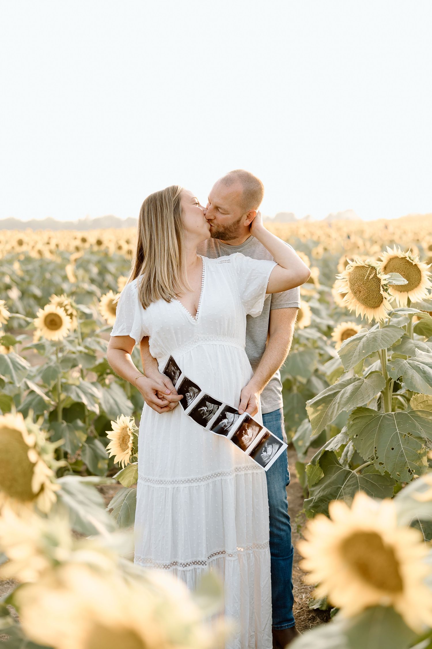 A romantic couple embraces in a sunflower field while holding ultrasound photos during a sunset photoshoot.