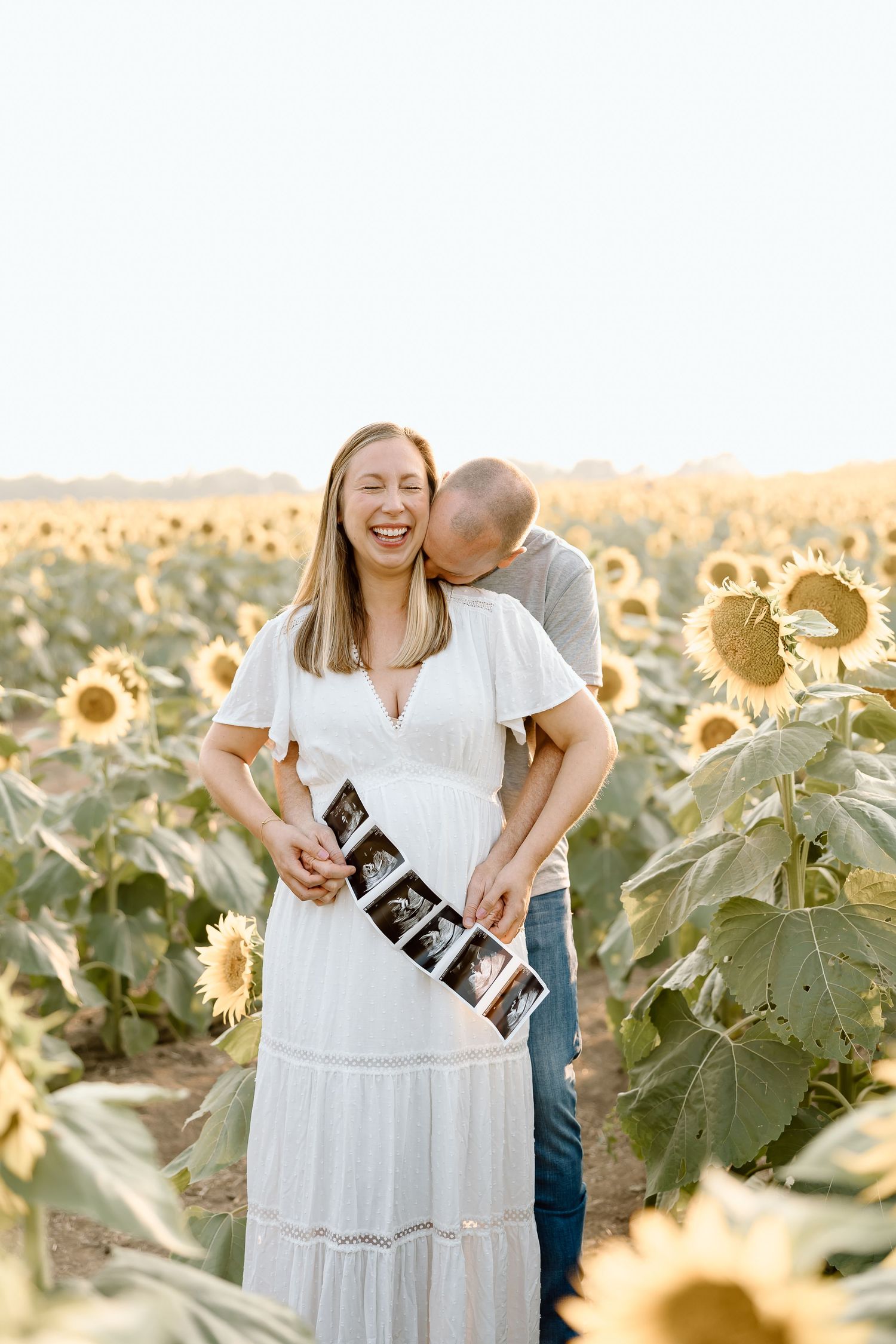 A couple embraces while holding ultrasound photos amidst a golden sunflower field during sunset.