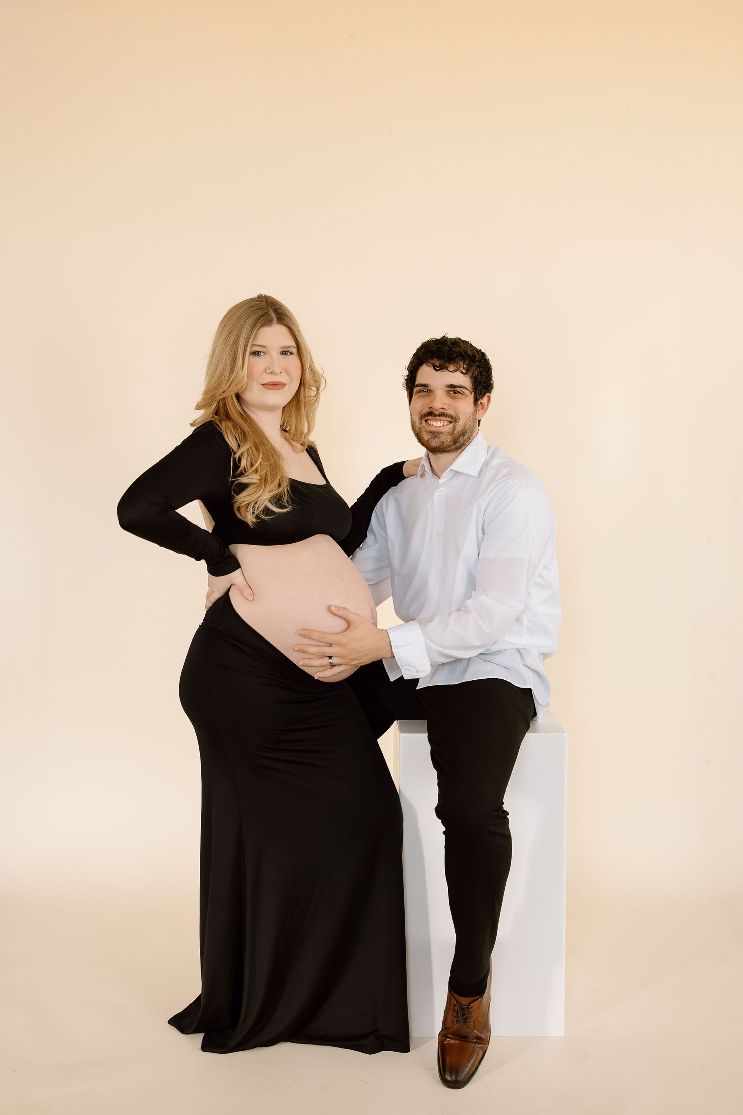 A couple poses together for a maternity photo series in black and white outfits against a beige background.