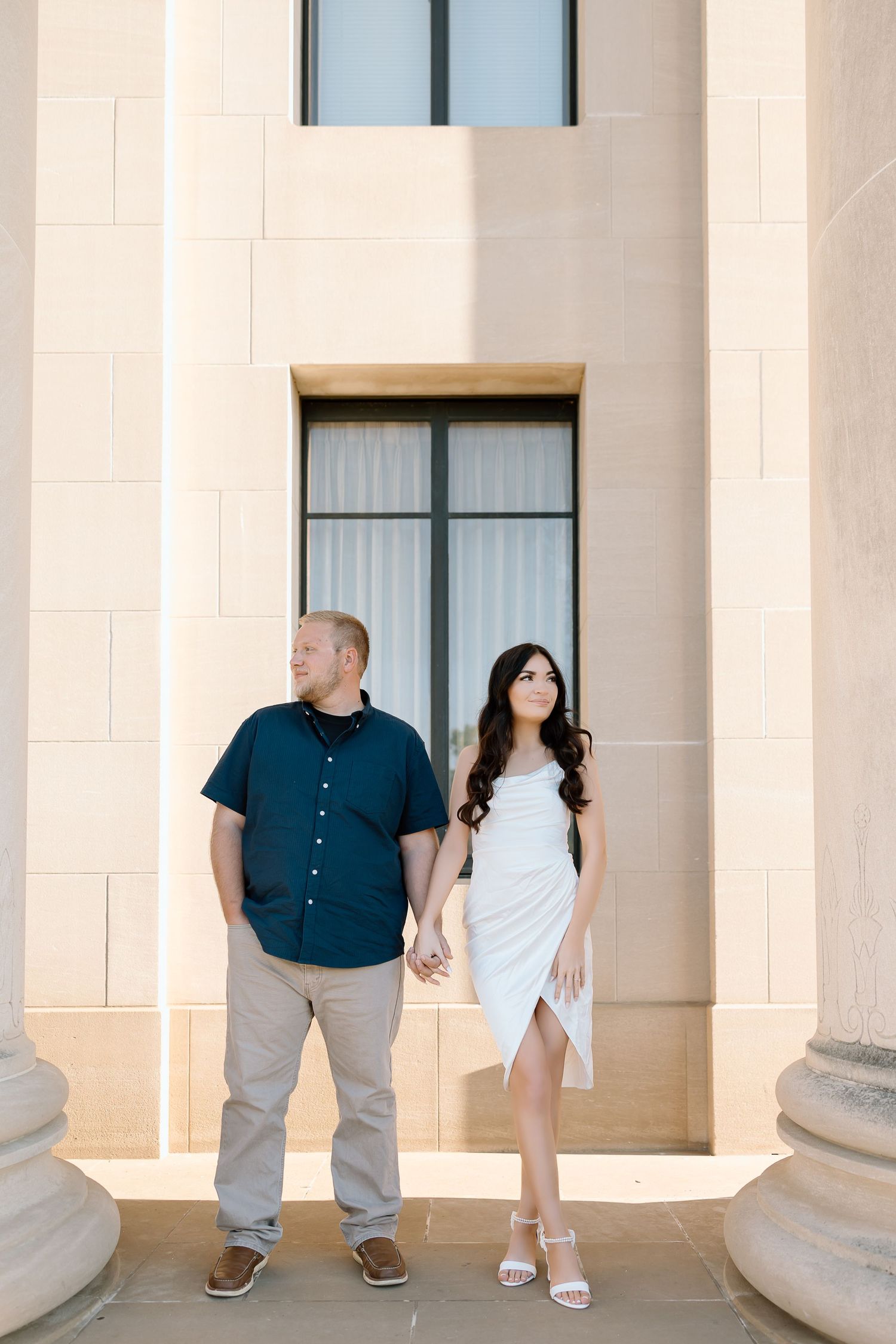 A couple stands between white columns in an elegant architectural setting during a photoshoot.