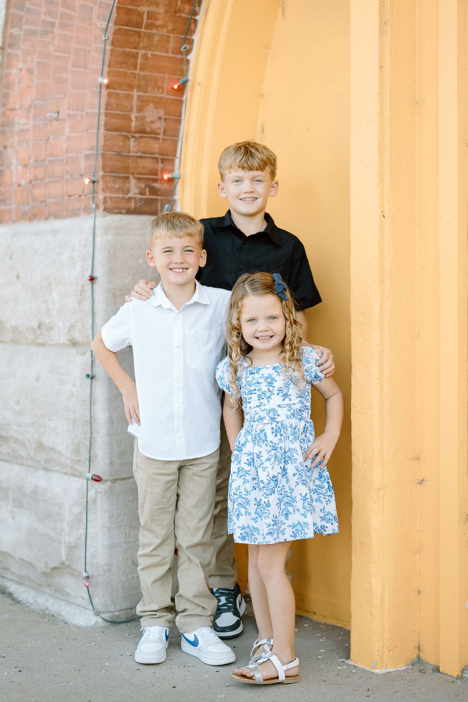 Three children stand together against a yellow wall with exposed brick, wearing casual clothing for a portrait.