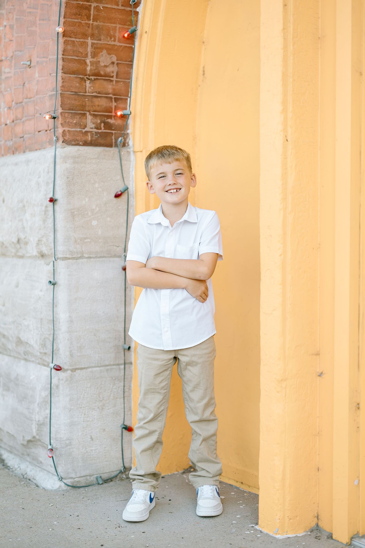 A child in a white polo shirt and khaki pants stands smiling against an orange and brick wall.