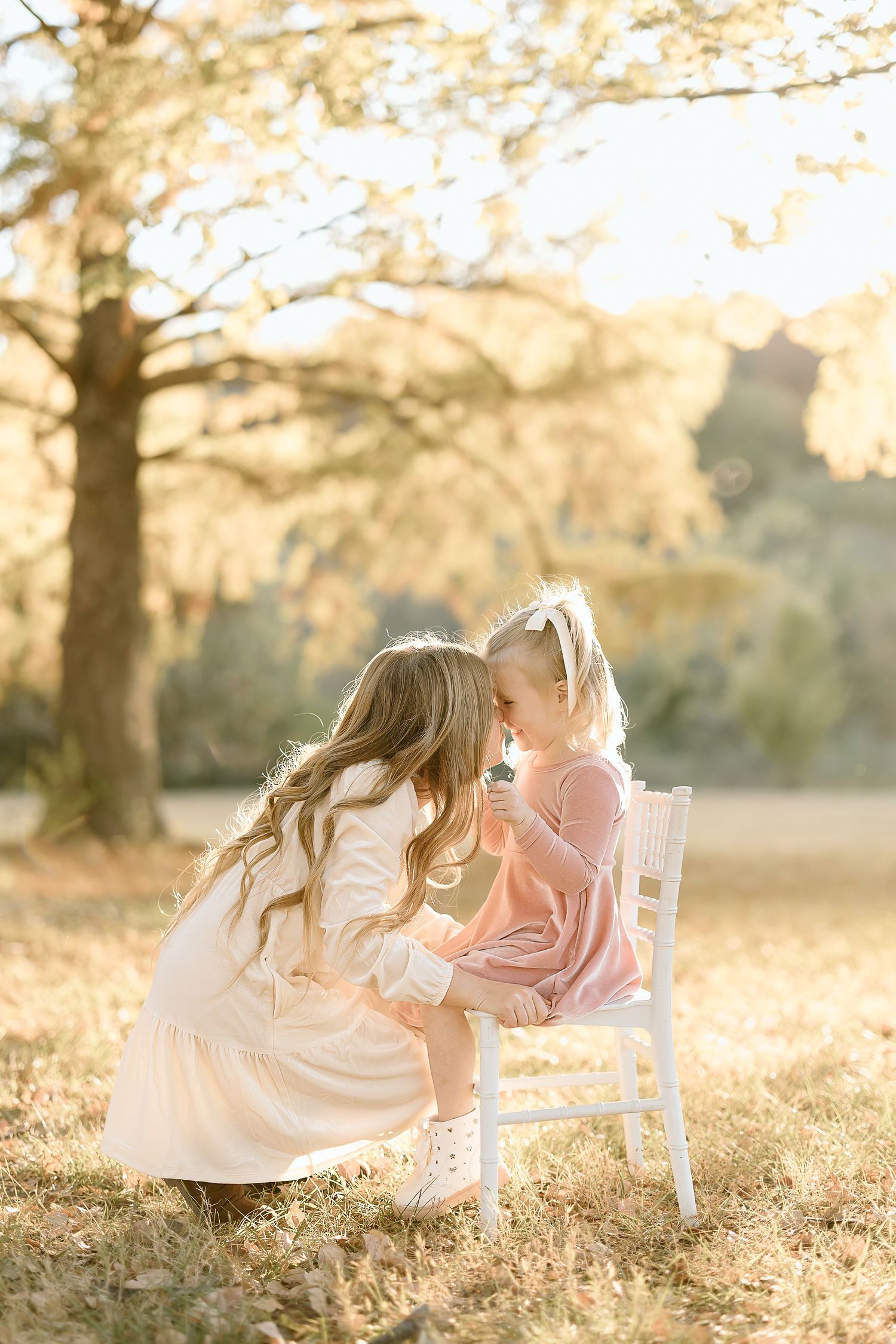 Two young girls in white dresses play together on a white chair in a sunlit field during golden hour.