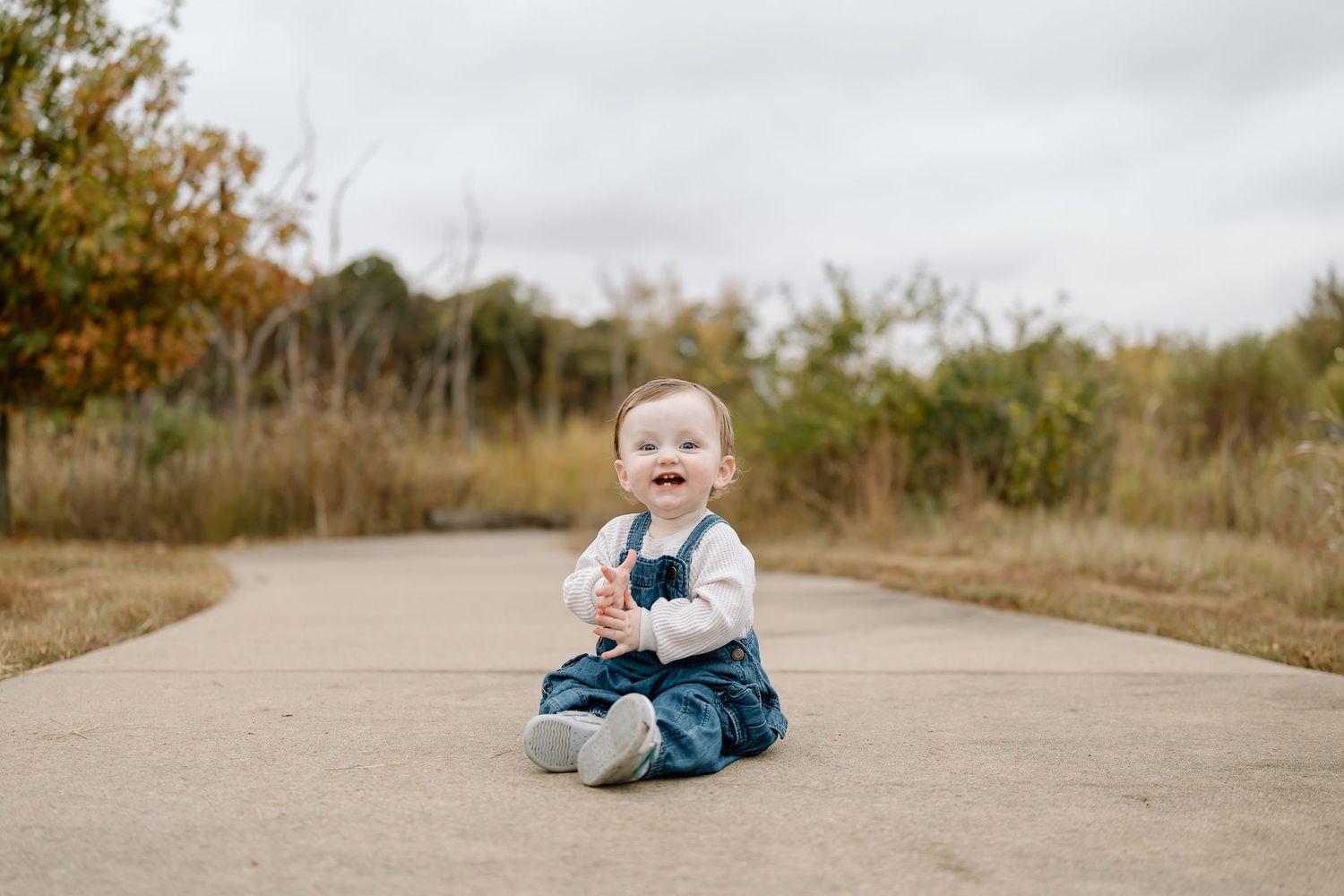 Small child wearing denim overalls sits and laughs on a winding concrete path during autumn.