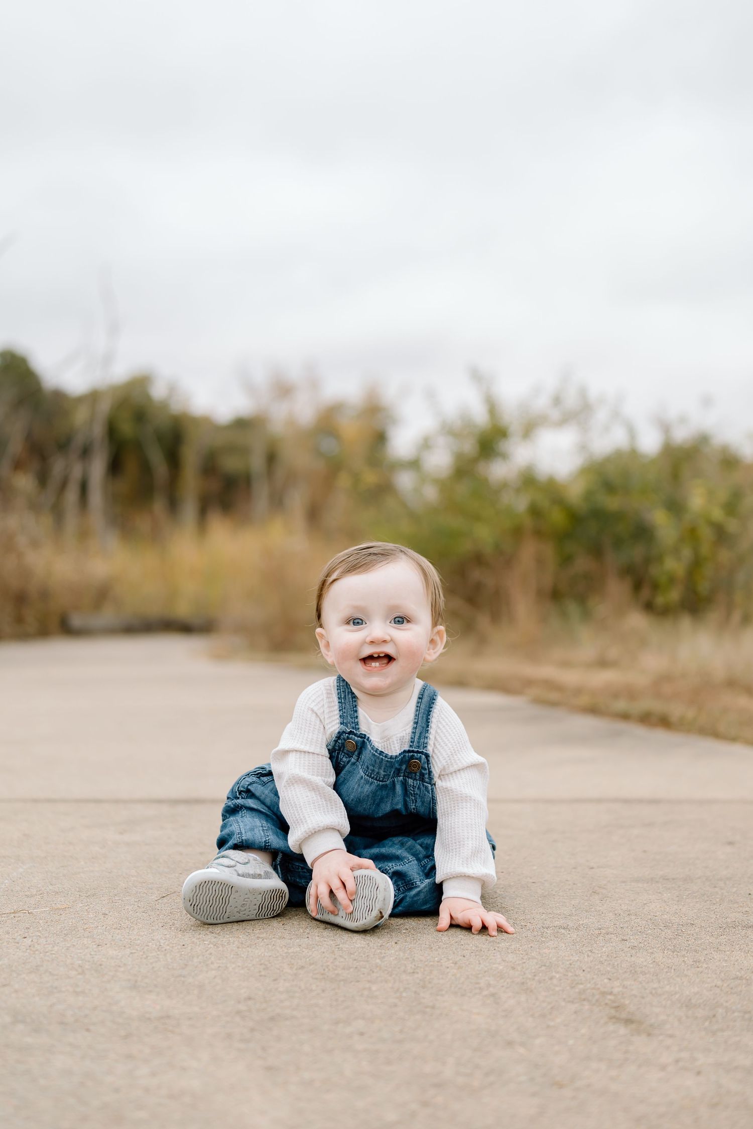 Baby in blue overalls and white shirt crawls and plays on concrete path during outdoor photo session.