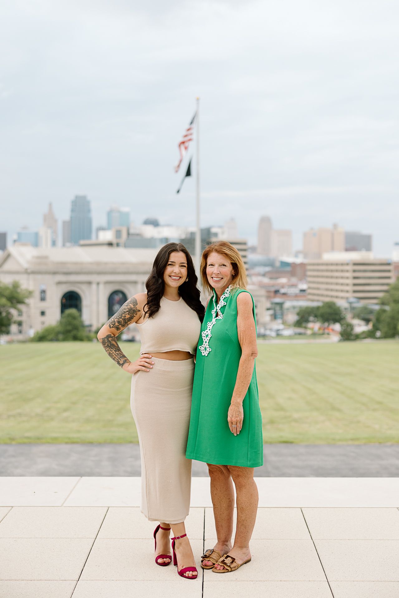 Two friends in beige and green dresses pose together on urban rooftop with city skyline.