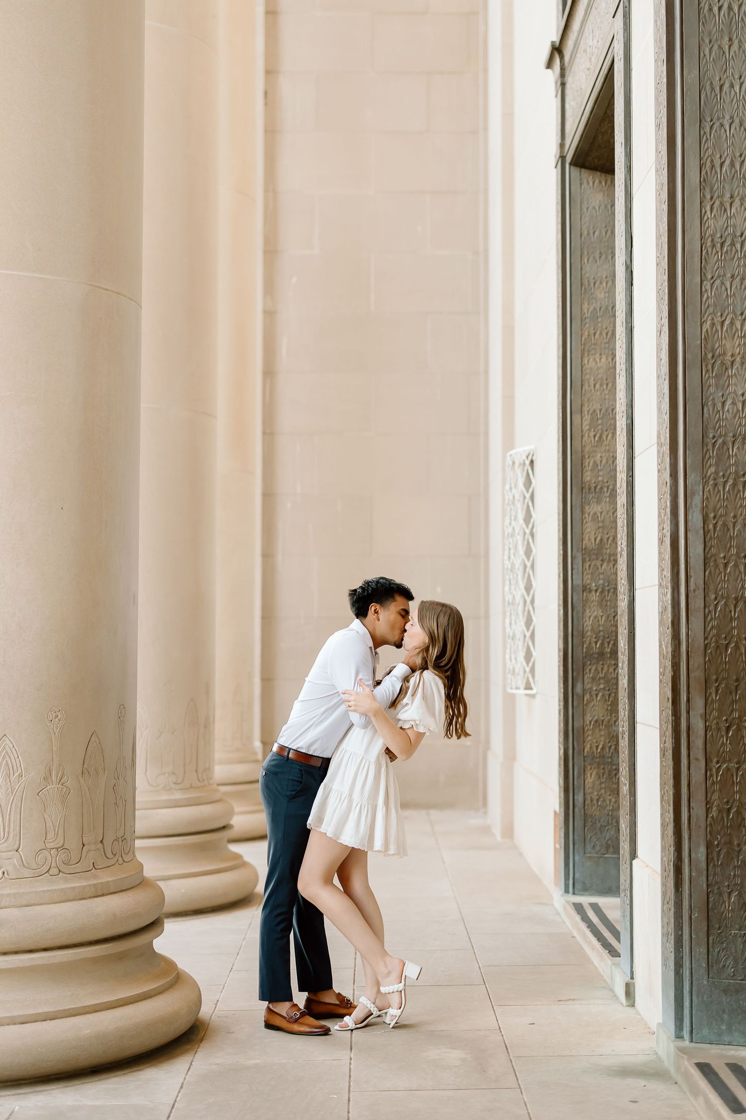 A romantic couple shares tender moments in an elegant colonnade with white stone pillars and natural light.