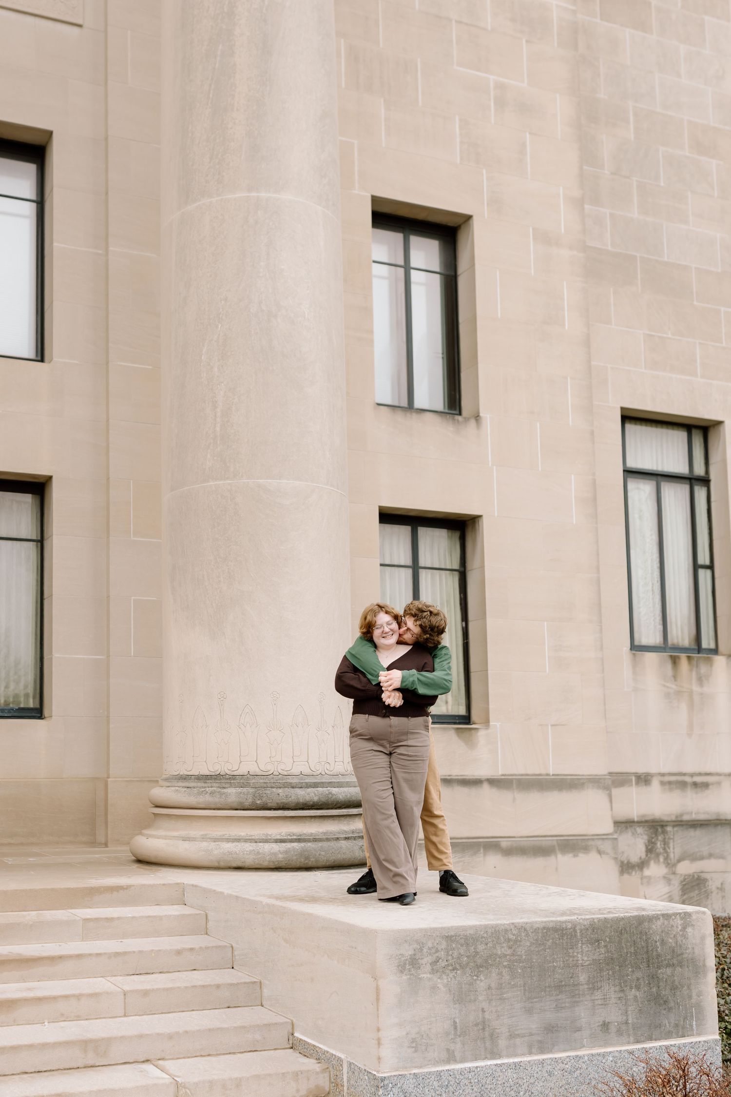 A couple shares a romantic kiss on concrete steps outside a stone building with windows.